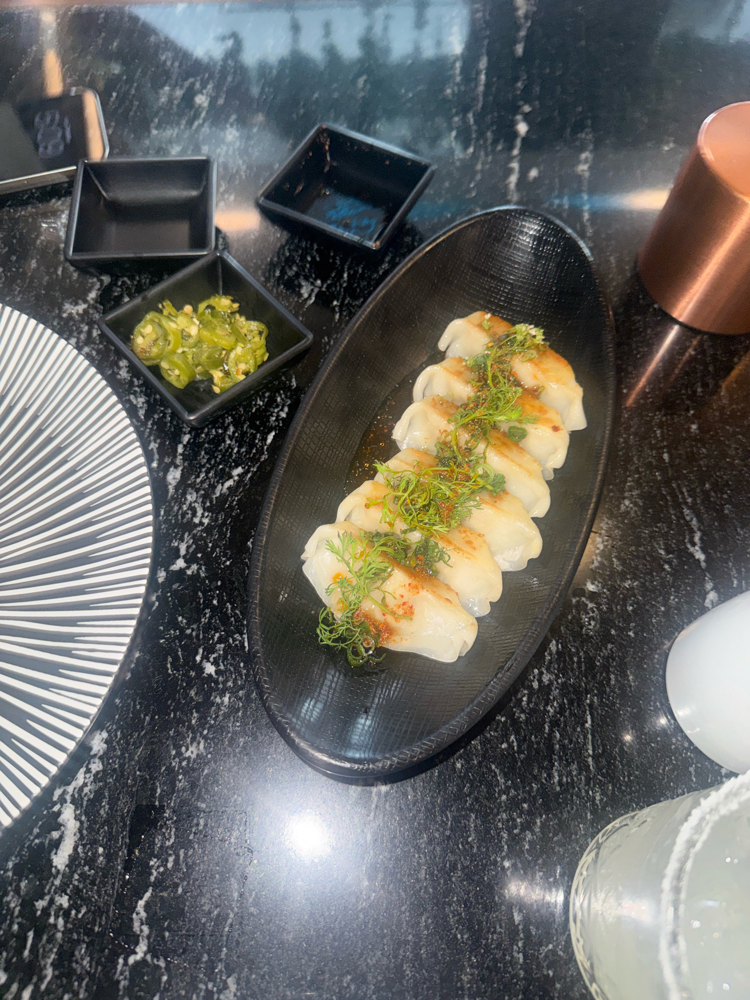A close-up, overhead shot showcases a dimly lit restaurant table, possibly in Puebla, Mexico, featuring a dark, speckled granite surface. In the foreground, to the left, a portion of a white and black patterned plate is visible. To the right of this, a small black square dish contains sliced green jalapeños. Behind this, another empty black square dish is positioned. Central to the image is an oval black plate holding six pan-fried dumplings, garnished with fresh green herbs and a dusting of red chili flakes. To the right of the dumplings, a copper-colored metal object, possibly a cup or container, stands upright. Further to the right, a salt-rimmed glass containing a light-colored beverage is present. A white, spherical object, likely a light fixture or decorative item, is also visible. The overall ambiance suggests a modern dining establishment.