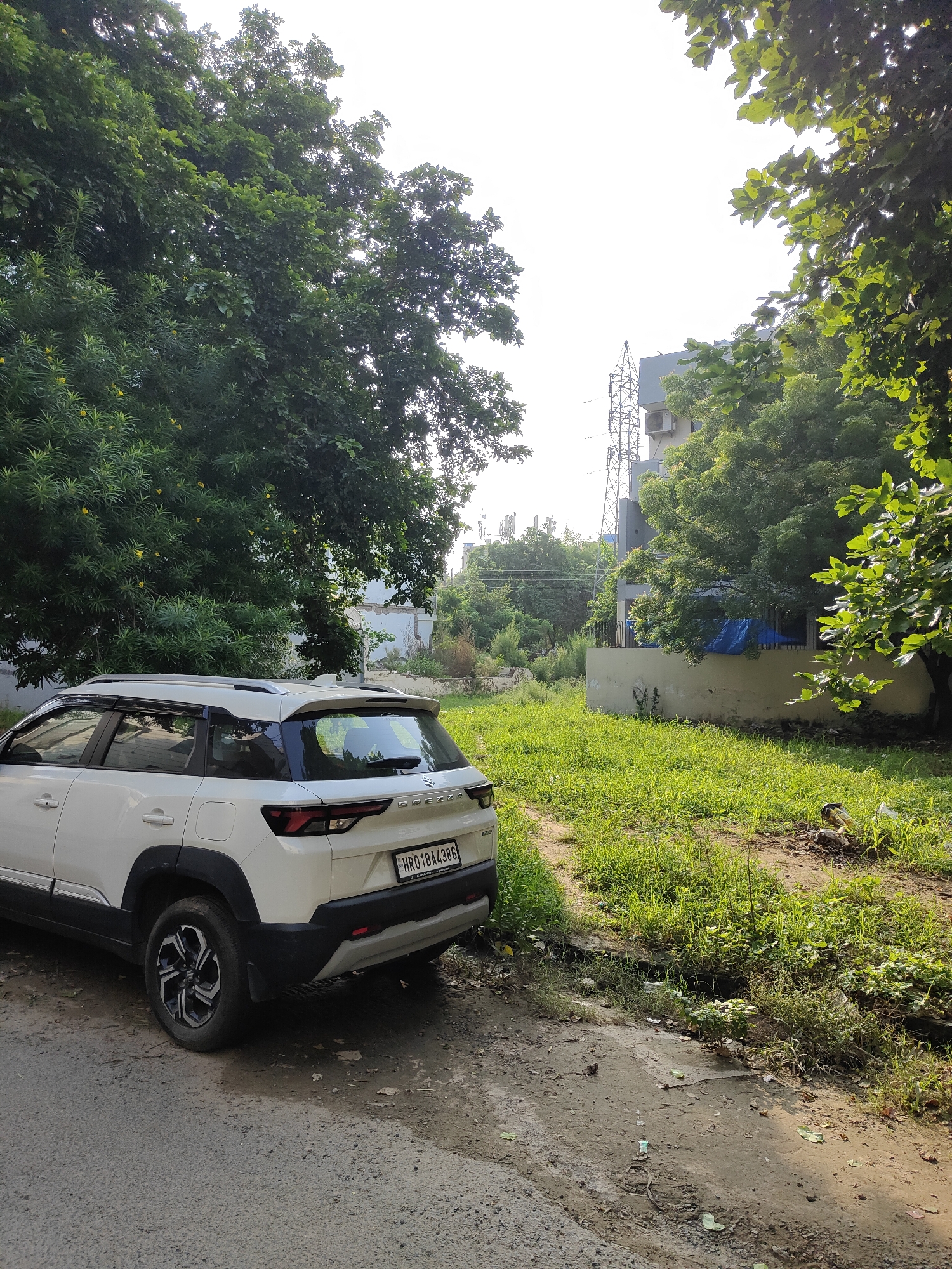 A white SUV, with a license plate that reads "HR01BA4386", is parked on the side of a road in Gurgaon, India. The car is positioned at an angle, with its rear end closer to the foreground. In the background, there is a small, overgrown plot of land with tall green grass and some scattered debris. A low, beige-colored wall runs along the edge of the plot, and behind it, a building with multiple stories can be seen, along with a tall lattice-like tower structure. Lush green trees surround the area, with some branches extending into the frame from the left and right sides, partially obscuring the sky. The sky itself is bright and hazy, suggesting it might be daytime. The road in the foreground appears to be paved with asphalt and shows signs of wear and tear.