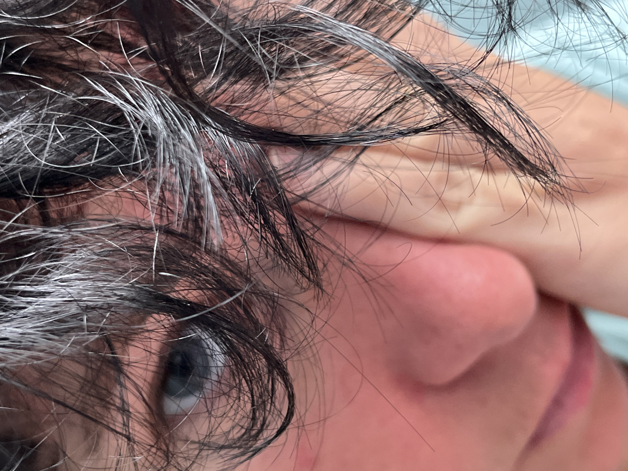 A close-up, angled shot focuses on the left side of a person's face, obscured by dark, curly hair. The person's eye, with a blue iris and dark pupil, is visible through the strands of hair. The skin of the face is pink, and the person's left hand is touching their cheek. The background is a blurred light blue. The image was taken in the village of Rkhetvi, Dehali Community, Georgia.