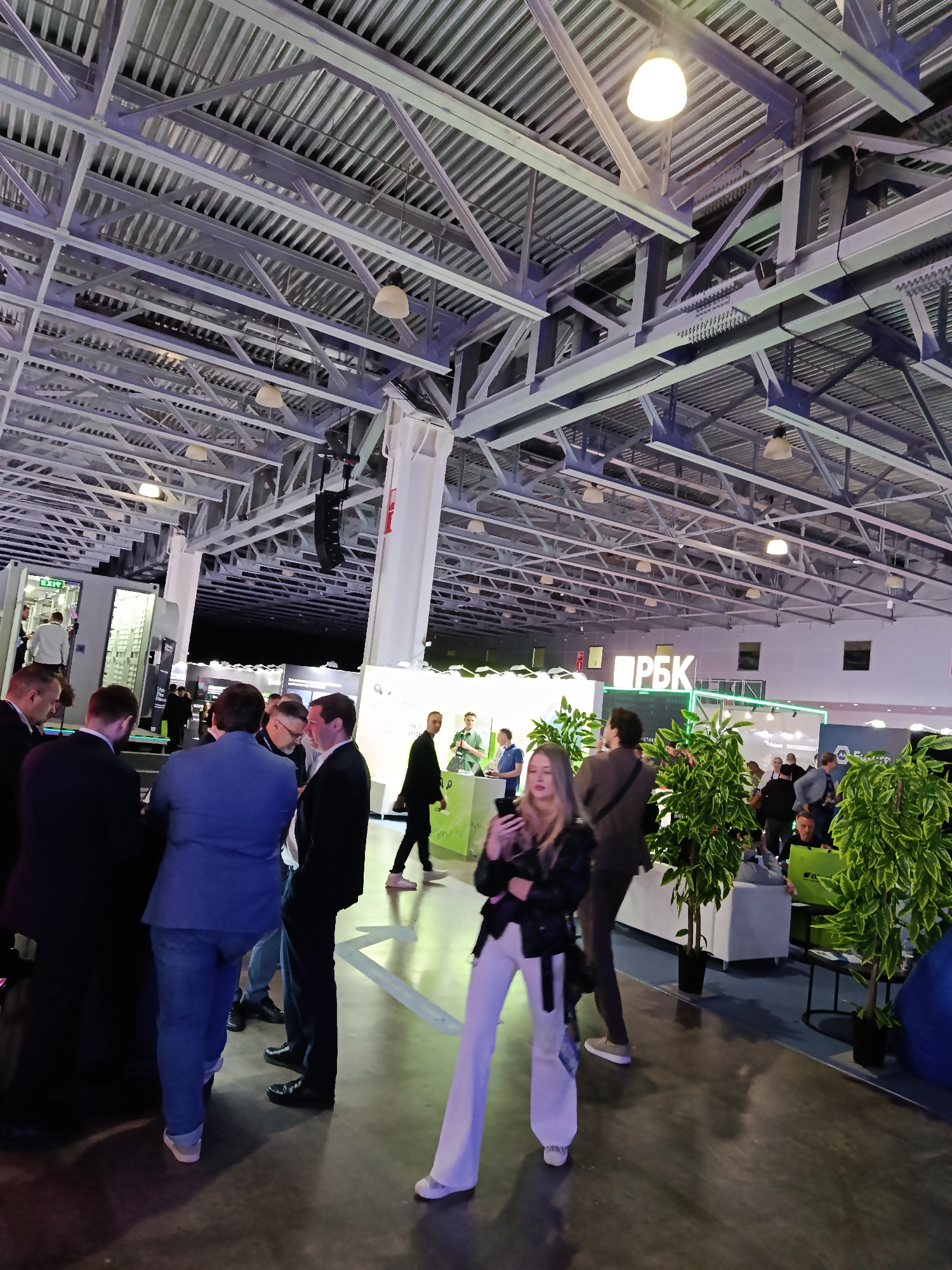 The image shows an indoor event or conference taking place in what appears to be a large hall, possibly an exhibition center. The location is likely Khimki, Russia, based on the text "РБК" displayed prominently on a stand. The hall has a high ceiling with a complex network of metal beams and overhead lighting fixtures. Groups of people are scattered throughout the space, some standing and conversing, others walking between the stands. A woman in white pants and a black jacket stands in the foreground, looking at her phone. There are potted plants near the "РБК" stand, and the scene suggests a business or technology-related event.