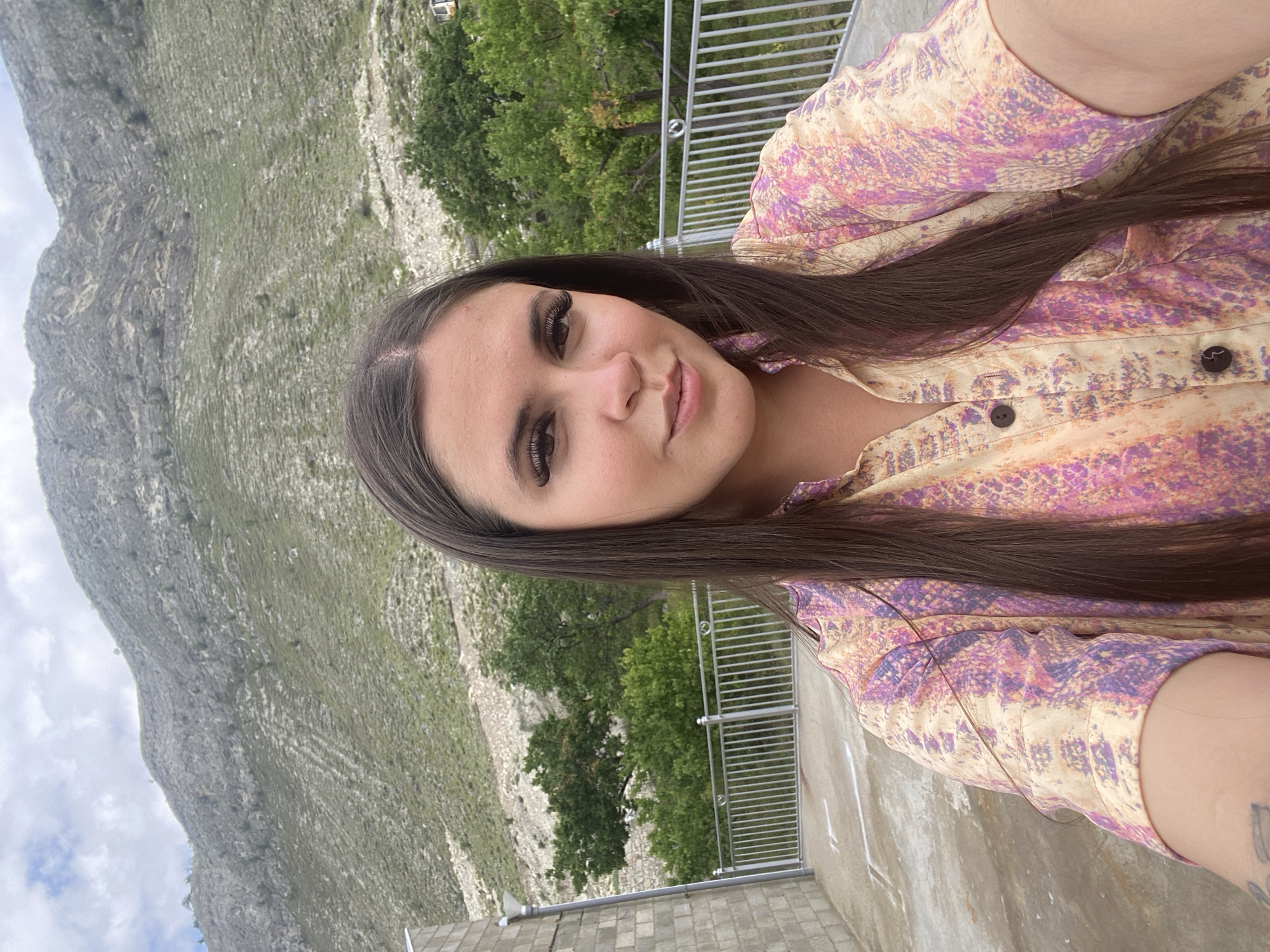 A young woman with long, dark hair and dramatic false eyelashes takes a selfie outdoors. She is wearing a short-sleeved button-up shirt with a colorful, abstract pattern in shades of pink, orange, and purple. Behind her, a large, barren, rocky mountain slopes upward, sparsely covered with green vegetation and dotted with a few trees. A metal railing runs horizontally across the middle ground. To the left, a portion of a textured, grey brick building is visible, and the ground appears to be a concrete surface with white lines painted on it. The sky above is cloudy. The location is identified as Chalda, Russia, though this specific scene does not contain any discernible urban elements that would confirm it is within a city.