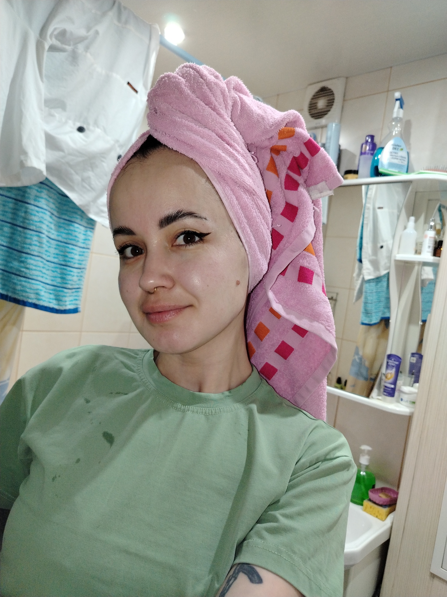 A young woman stands in a bathroom in Chaikovsky, Russia. She is wearing a light green t-shirt with some spots on it and has a pink towel wrapped around her head. She has dark eyeliner and a nose ring. Behind her, clothes are hanging on a rack, and bathroom supplies are visible on shelves and in a mirrored cabinet. A small ventilation fan is on the wall above the shelves. A tattoo is visible on her left arm.