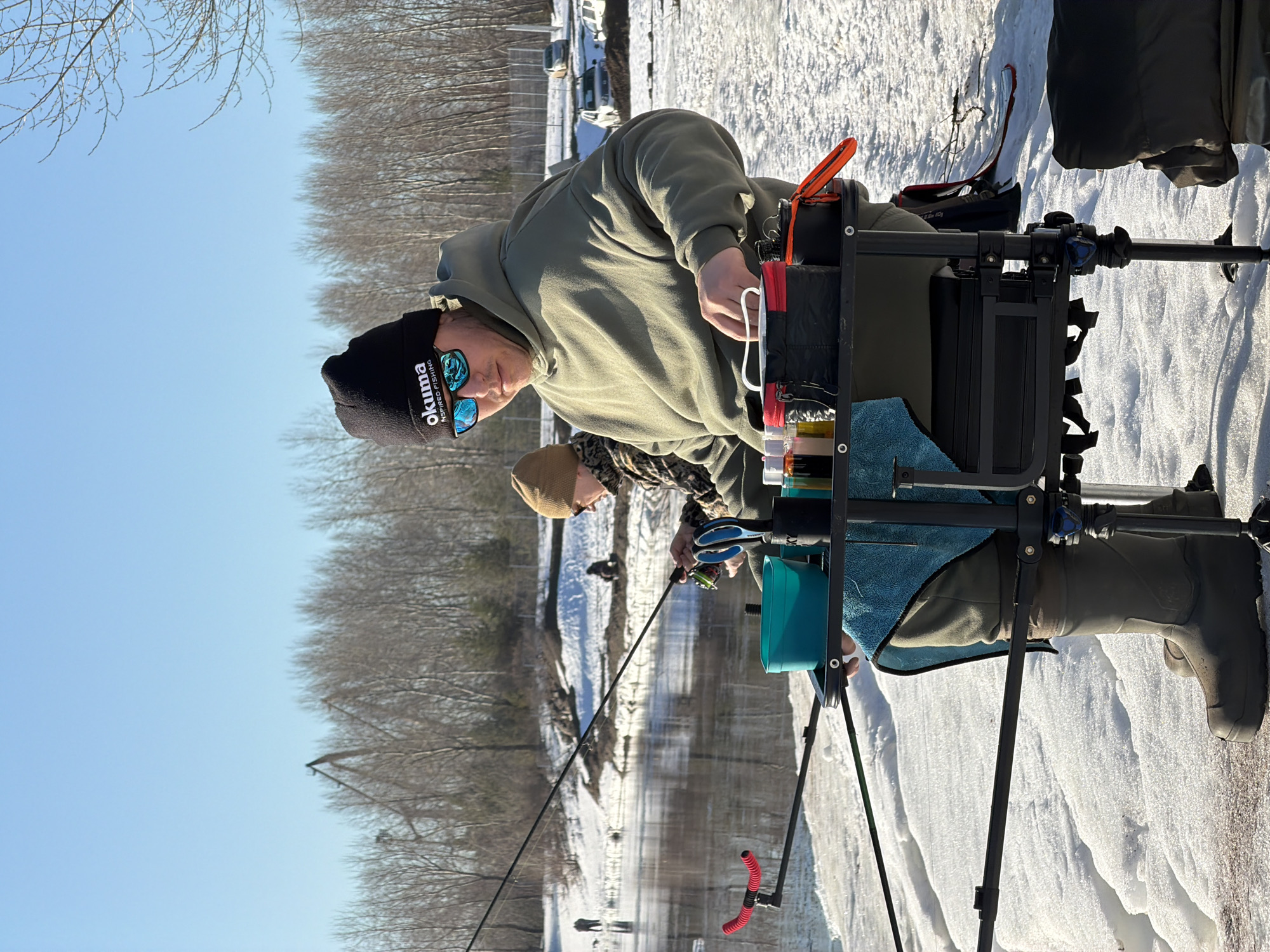Two individuals are ice fishing on what appears to be a frozen body of water. The foreground is dominated by a man in a black Okuma beanie, mirrored sunglasses, and a green hoodie, seated on an elevated fishing platform with various tools and accessories attached. He seems to be preparing his equipment. Behind him, another man in camouflage attire is actively fishing with a rod and reel. The surrounding environment is snow-covered, with bare trees visible in the background. A glimpse of buildings and vehicles indicates the proximity of a town or city. The location is identified as Noviy, Russia.
