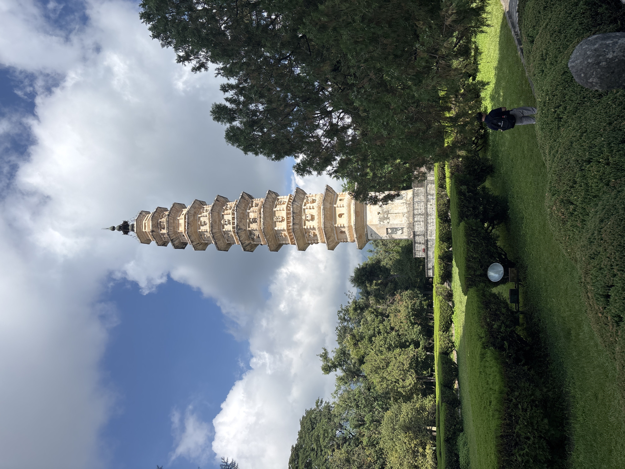 The image shows the Three Pagodas of Chongsheng Temple in Dali City, China, under a partly cloudy blue sky. The tallest of the three pagodas, which is the central and most prominent one, is visible in the background. It is an ancient Buddhist stupa, octagonal in shape and constructed from brick. It has multiple tiers, each decorated with intricate carvings and detailing, and is topped with a spire.
In the foreground and mid-ground, there is a well-maintained garden with lush green grass and meticulously trimmed hedges. A solitary person, viewed from behind, is walking on the grass in the garden, wearing a dark shirt and light-colored pants, and carrying a backpack. Trees, including a large evergreen with dense foliage, frame the scene. A round, reflective object, possibly a mirror or a sign, is placed on a low structure within the garden. The overall impression is of a serene and historically significant site.
