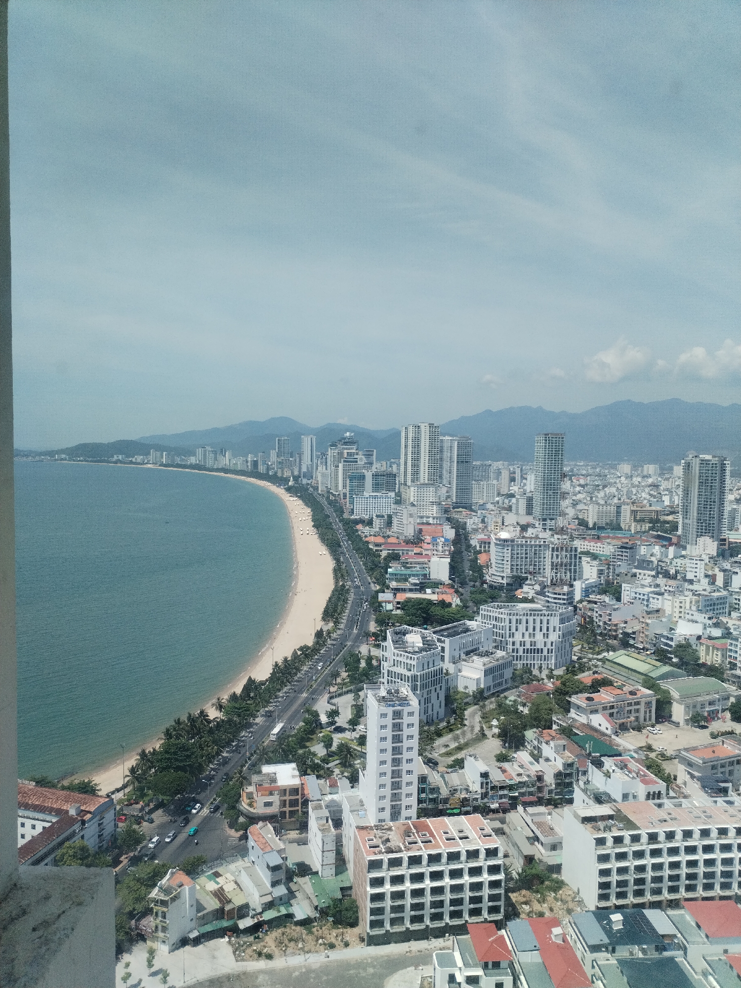 This is a high-angle, full shot of Nha Trang, Vietnam, focusing on a curved, sandy beach lined with palm trees and a coastal road. The ocean, a calm turquoise, meets the shore, and a wide expanse of city buildings, including numerous high-rise structures and shorter buildings, stretches inland. In the distance, a range of hazy, blue mountains forms the backdrop under a light, cloudy sky. The scene appears to be captured during daylight, suggesting a warm climate. The city is densely populated with a mix of modern and older architecture. There are no visible people or specific actions, creating a still, observational portrayal of the cityscape and its natural surroundings.