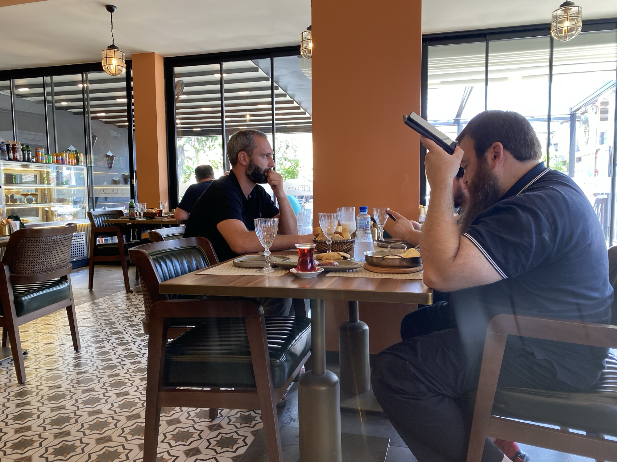 In a brightly lit restaurant in Sokhumi, Abkhazia, two men sit at a wooden table. The man on the left, with a beard and short dark hair, wears a black polo shirt and is leaning forward, resting his chin on his hand. The man on the right, who is larger and has a full beard, wears a dark blue polo shirt and is holding a tablet, looking down at it. In front of them, the table is set with glasses, a small cup of tea, a basket of bread, and plates of food. The restaurant's interior features orange walls, large windows with a view of the outdoors, and a distinctive patterned floor. Two wooden chairs with dark green upholstery are visible, one in the foreground and another to the left.