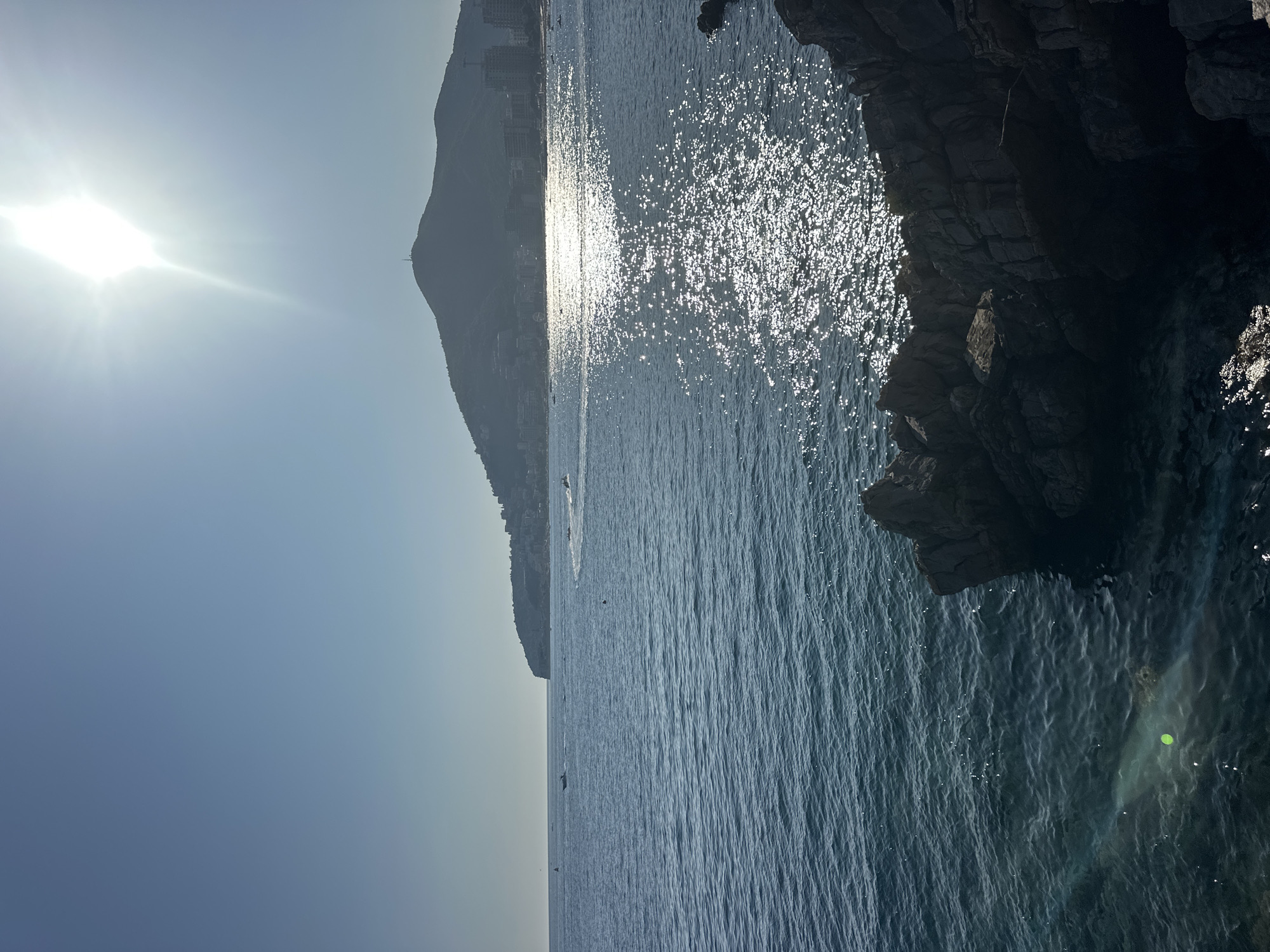 The image depicts a scenic coastal view of Budva, Montenegro, likely captured from a rocky outcrop. The foreground features a cluster of dark, rugged rocks that lead into the clear, deep blue sea. Sunlight glints off the water's surface, creating a shimmering path that extends towards a distant, hilly coastline. This coastline is dotted with buildings and shows signs of development, including what appear to be hotels or apartment complexes.
In the middle ground, several small boats are visible on the water. One boat appears to be a speed boat or jet ski, leaving a white wake as it moves. Other, smaller vessels are further out, possibly fishing boats or pleasure craft. The sky is a clear, pale blue, with the sun positioned high and slightly to the right, its bright glare creating a lens flare effect in the image. The overall atmosphere is one of a sunny, active day at the seaside.