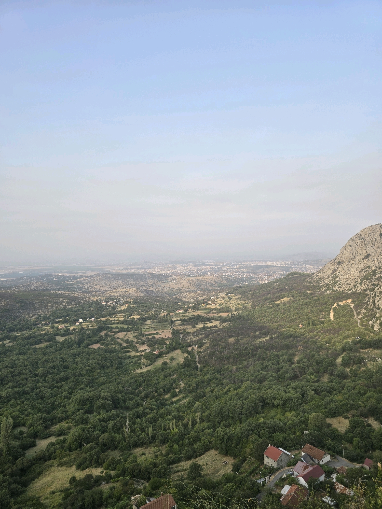 The image shows a panoramic view of a mountainous landscape with a town or city in the distance. The foreground is dominated by lush green trees and vegetation, with a few scattered houses with red roofs visible on the slopes. A prominent rocky mountain rises on the right side of the image. The background features a hazy view of a sprawling urban area, likely a city, extending across the horizon. The sky is a pale blue with some light clouds. The scene appears to be captured from an elevated vantage point, looking down over the valley and the distant city.