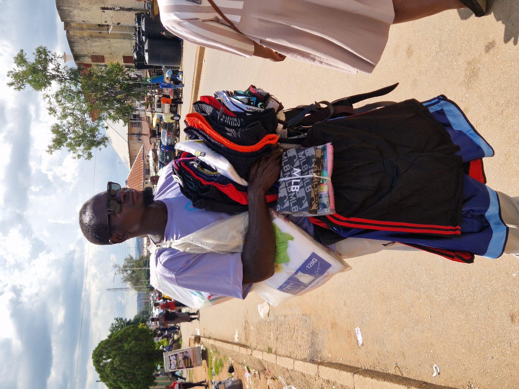 A dark-skinned man wearing sunglasses and a purple t-shirt walks along a paved road in Jos, Nigeria, carrying a load of folded clothes over his shoulder and in his arms. The man has dark hair and a beard. The clothes are a mix of shorts and t-shirts, some of which are colorful. He appears to be a street vendor. In the background, there are buildings, parked cars, and other pedestrians. The sky is cloudy.