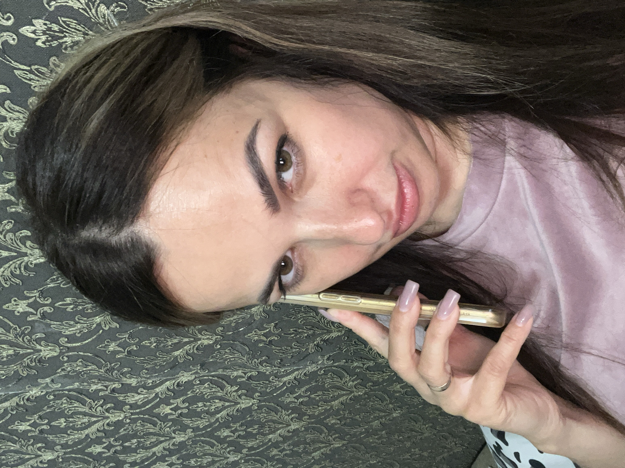 A close-up, eye-level, indoor selfie shows a woman with long, dark brown hair, and fair skin, holding a gold smartphone to her right ear with her right hand. She is wearing a pale pink, possibly velvet, crewneck shirt. Her left hand is partially visible at the bottom of the frame, with long, light pink fingernails. The background consists of dark grey wallpaper with a repeating ornate, damask-like pattern in a lighter gold or green hue. The lighting appears to be from above and slightly to the left, casting subtle shadows on her face. The woman's gaze is directed slightly off-camera to her left.