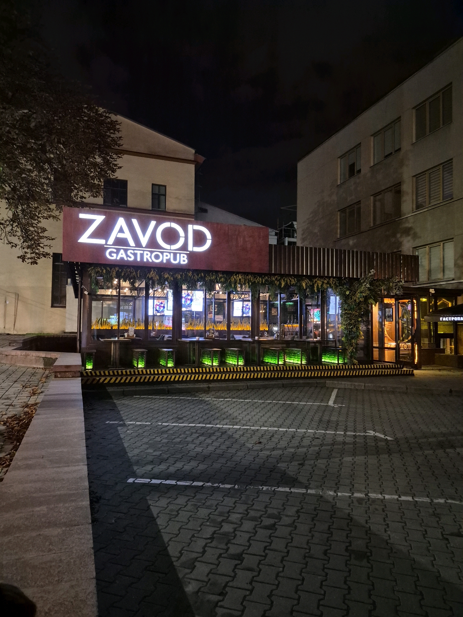 The image depicts the exterior of a gastropub named "ZAVOD" in Minsk, Belarus, at night. The gastropub features a modern design with a glass-enclosed seating area illuminated by green lighting and decorative foliage. The signage prominently displays "ZAVOD GASTROPUB" in large white letters against a dark red background. The surrounding environment includes a cobblestone parking lot marked with white lines, a sidewalk with fallen leaves, and adjacent buildings that appear to be apartments or commercial spaces. No people are visible in the frame.