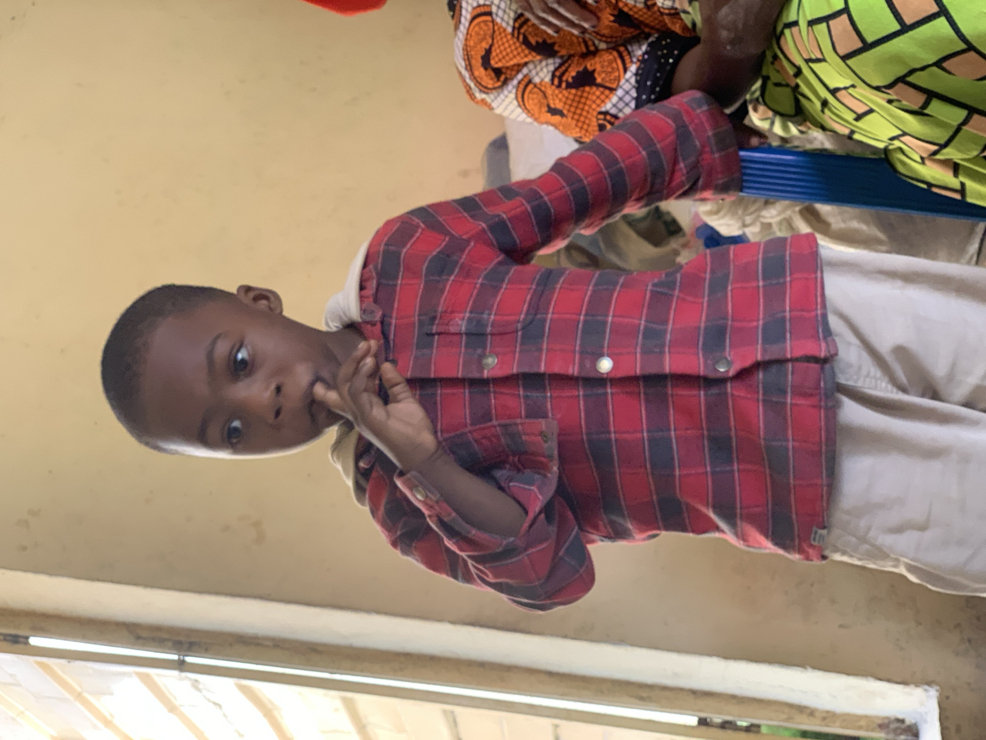 The image shows a young Black boy standing in what appears to be an indoor setting with a window on the left. He is wearing a red and black plaid shirt with a light-colored hood underneath and beige pants. His right hand is near his mouth, with his finger touching his lip. The background is a plain, light-colored wall. To the right of the boy, a portion of another person is visible, wearing a vibrantly patterned garment with orange, green, and black colors. The overall lighting suggests it is daytime. The image does not contain any explicit indicators of its specific location within Jos, Nigeria.