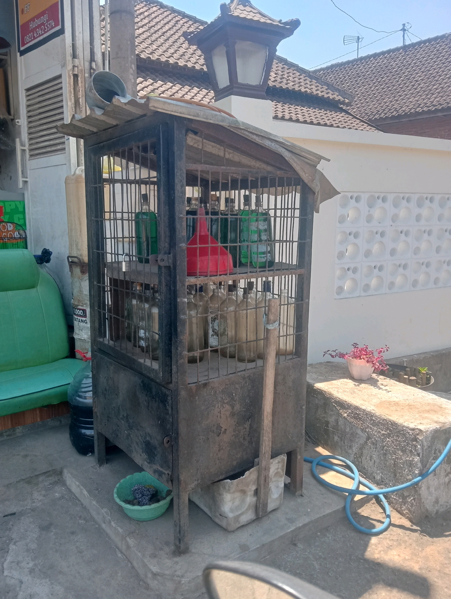This is a medium-shot, eye-level, outdoor photograph taken in Jatisari, Indonesia. The scene depicts a cage-like structure containing several bottles of liquid, likely fuel, and a large red funnel. The cage is made of metal bars and has a weathered, rustic appearance. It is situated on a concrete surface, with a blue hose coiled nearby. In the background, there is a white building with distinctive patterned ventilation blocks and a tiled roof. To the left of the cage, a green car seat is visible, and a large plastic water container is behind it. The lighting suggests it is daytime, with a clear blue sky overhead. The overall impression is of a roadside stall or a small, informal fuel dispensing point.