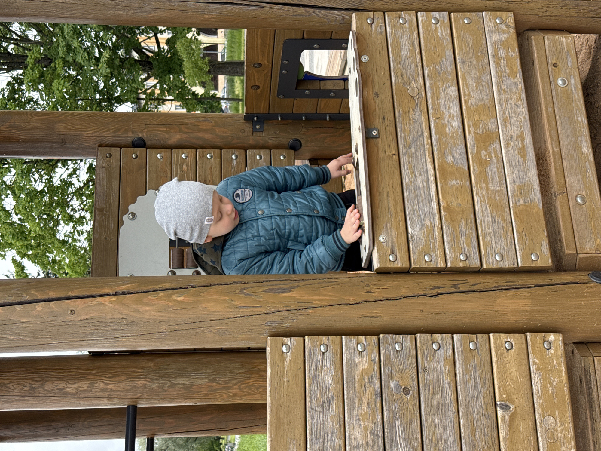 A child in a gray hat and a blue quilted jacket is playing on a wooden structure, possibly a playground. The wooden structure is made of planks secured with round metal fasteners. Trees and a building are visible in the background. The location may be Veliky Novgorod, Russia. The photo appears to be taken at an unusual angle.