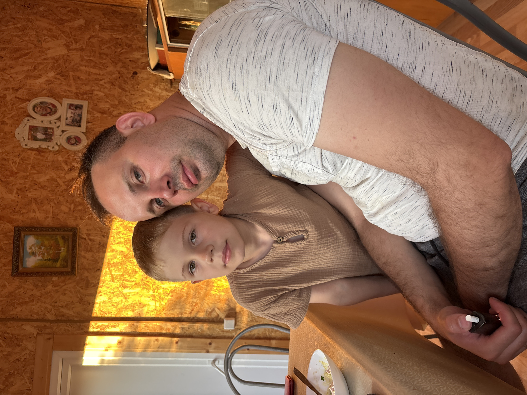 A man and a young boy are seated together, with the man's arm around the boy. The man is wearing a grey and white striped t-shirt and grey shorts, and he is holding a small black object with a white tip in his right hand. The boy is wearing a light brown, short-sleeved shirt. They are seated at a table covered with an orange tablecloth. Behind them, the wall is made of wood-paneled material. Sunlight is casting a strong, warm glow on the wall from a window on the left side of the image. A framed painting hangs on the wall above the boy's head, and a cluster of framed photographs is visible on the wall to the right of the painting. A chair with a metal frame is visible to the left of the table. The scene appears to be indoors in a rustic setting, possibly in Дунилово, Russia, given the architectural style and the presence of the sunlight through the window.