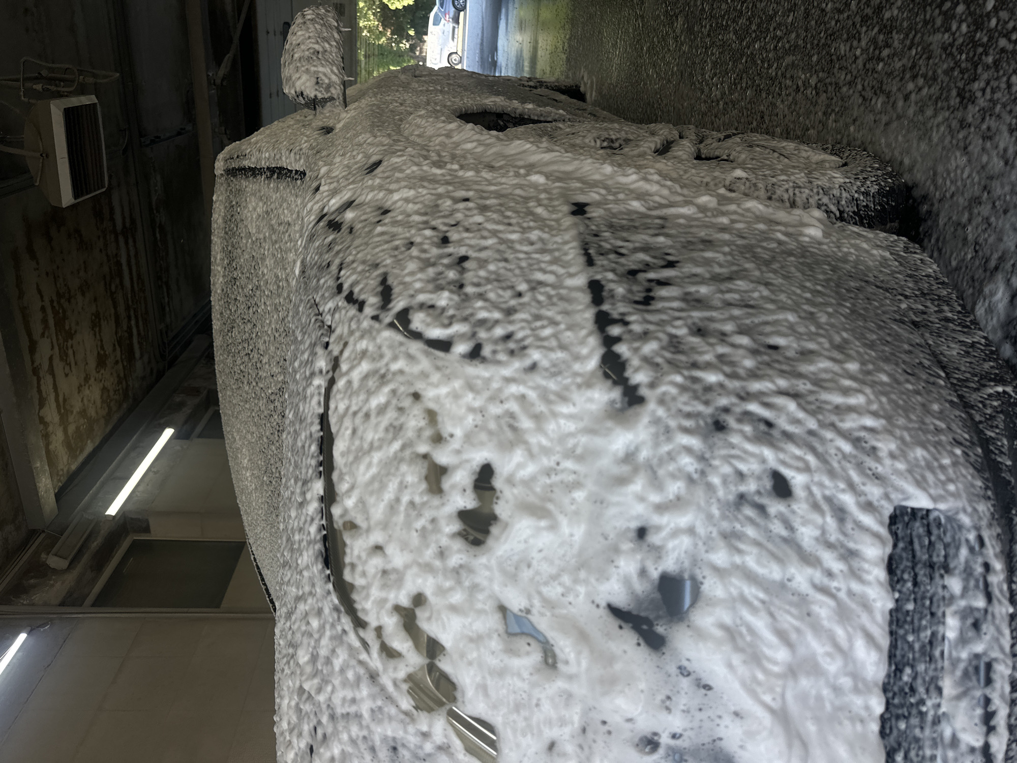 The image shows a car heavily covered in white foam at a car wash in Ternopil, Ukraine. The car is dark in color, with only glimpses visible beneath the foam. The car wash facility has exposed pipes, lights, and a wall mounted heater. Outside the car wash is a paved road, trees, and another vehicle.
