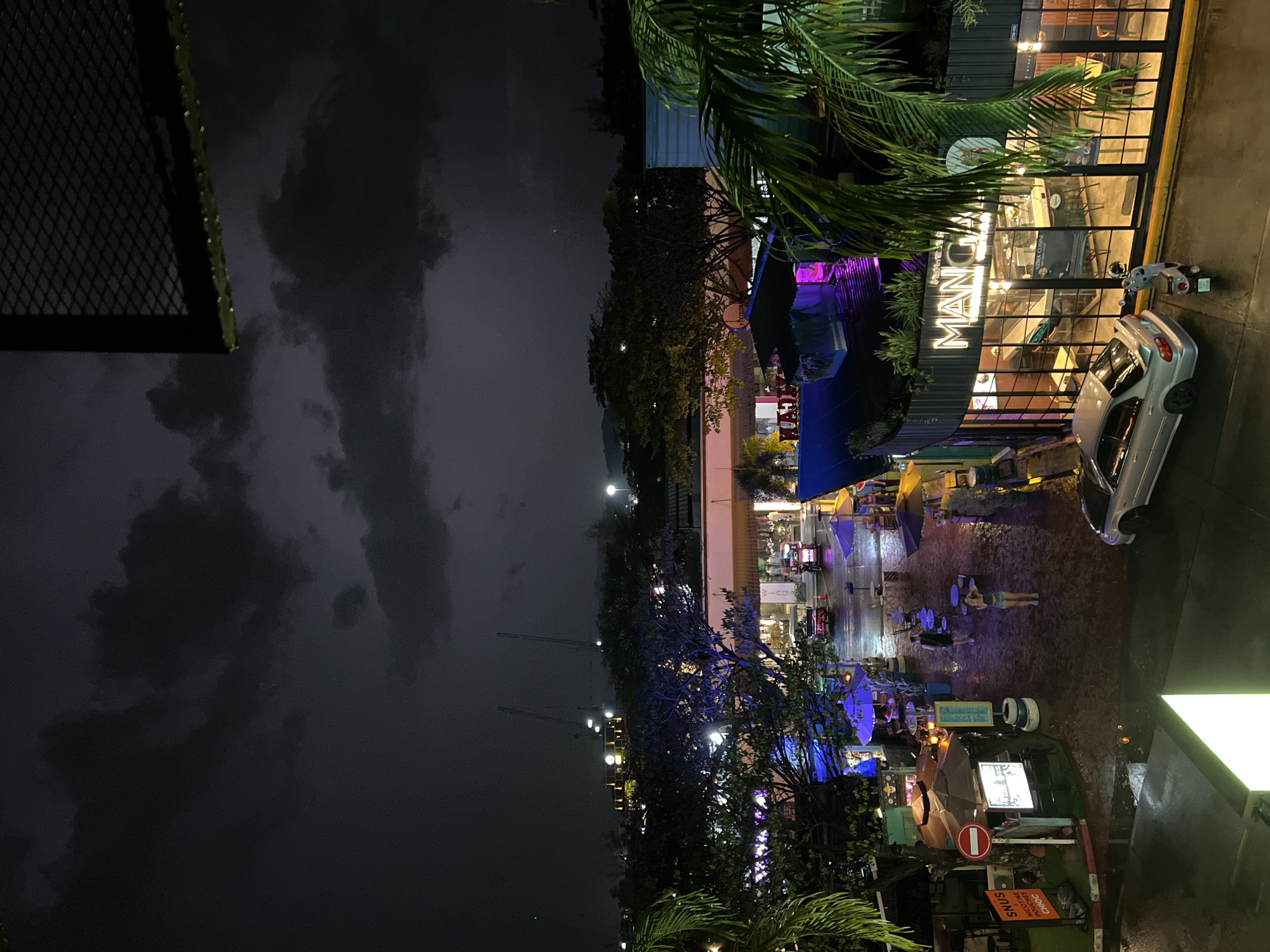 The image depicts an outdoor urban scene at night in an area with commercial establishments. The sky is dark with ominous clouds. A multi-story building with large windows and the illuminated name "MANCHO" is visible on the right side of the frame, with a silver car parked nearby. A sidewalk with several people and tables with umbrellas extends away from the building. The scene appears to be wet, possibly from rain. Street lights and colorful lights illuminate the area. In the foreground on the bottom right corner is a stop sign.