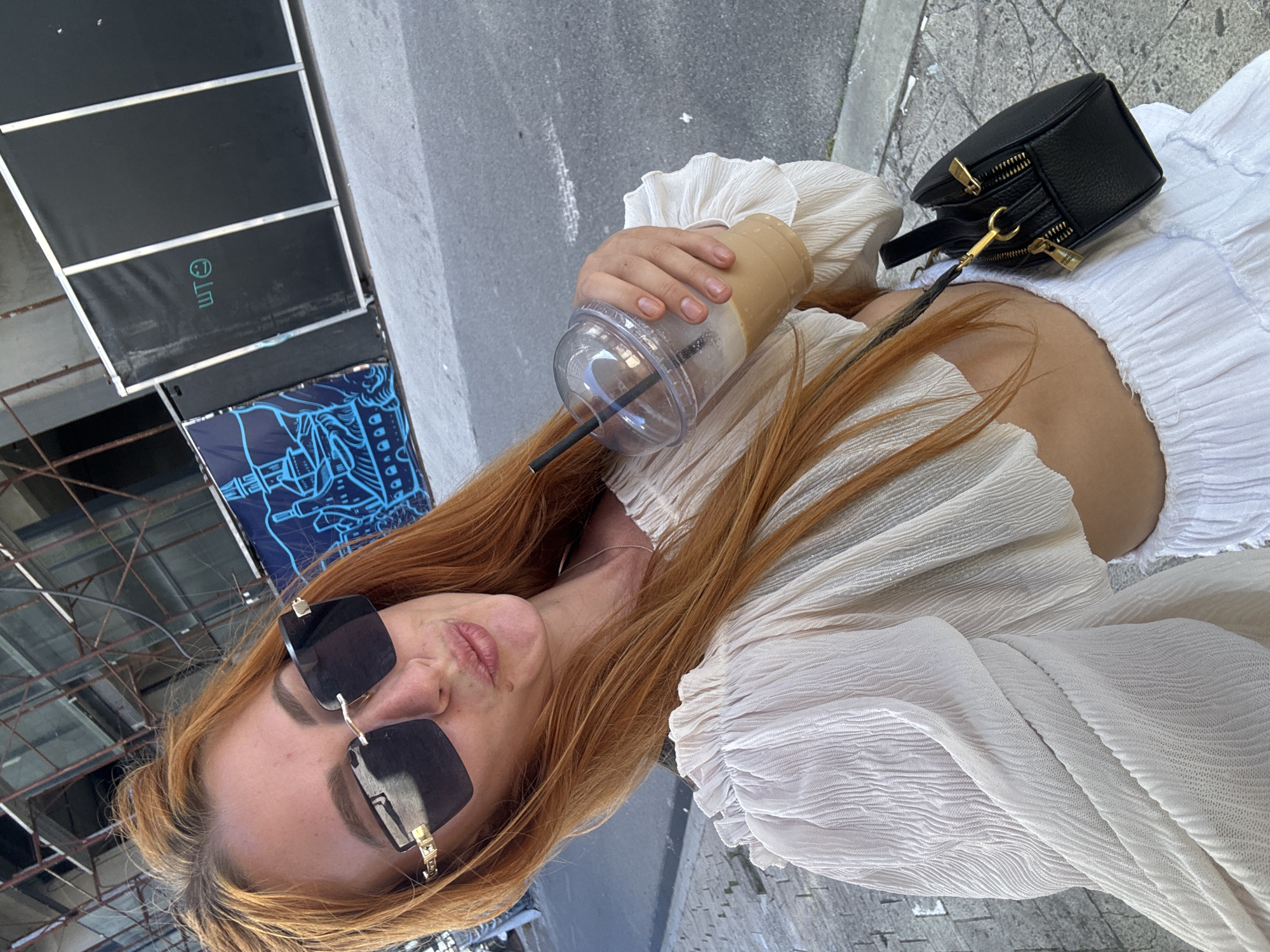 A woman with auburn hair is taking a selfie in Budva, Montenegro. She is wearing a white off-the-shoulder crop top with ruffled sleeves and a white ruffled skirt. She has on large black sunglasses and is holding a plastic cup with a beverage and straw. A small black handbag with gold hardware is hanging from her shoulder. In the background, there is construction scaffolding, a blue mural on a barrier, and a street.
