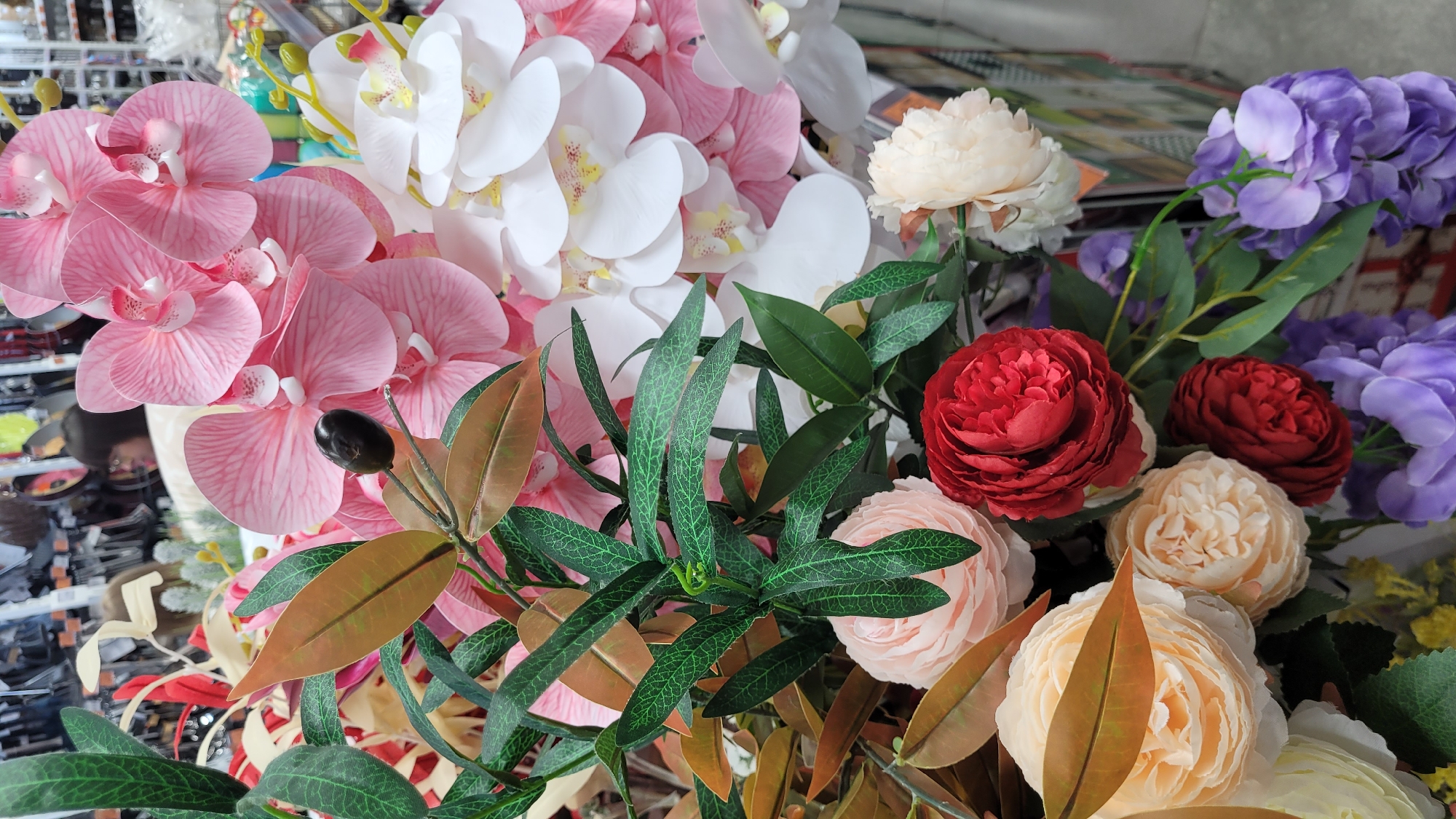 The image presents a collection of artificial flowers arranged in a manner suggestive of a display or bouquet. The flowers showcase a variety of colors and types, including pink and white orchids, peach-colored roses, deep red roses, and purple hydrangeas. Also present are artificial green leaves and branches, including some that mimic olive branches with simulated black olives. The background is indistinct, showing hints of a possible commercial environment, with a surface suggesting a table or display counter. The location within the city of Unknown, Unknown, cannot be determined from the provided image.