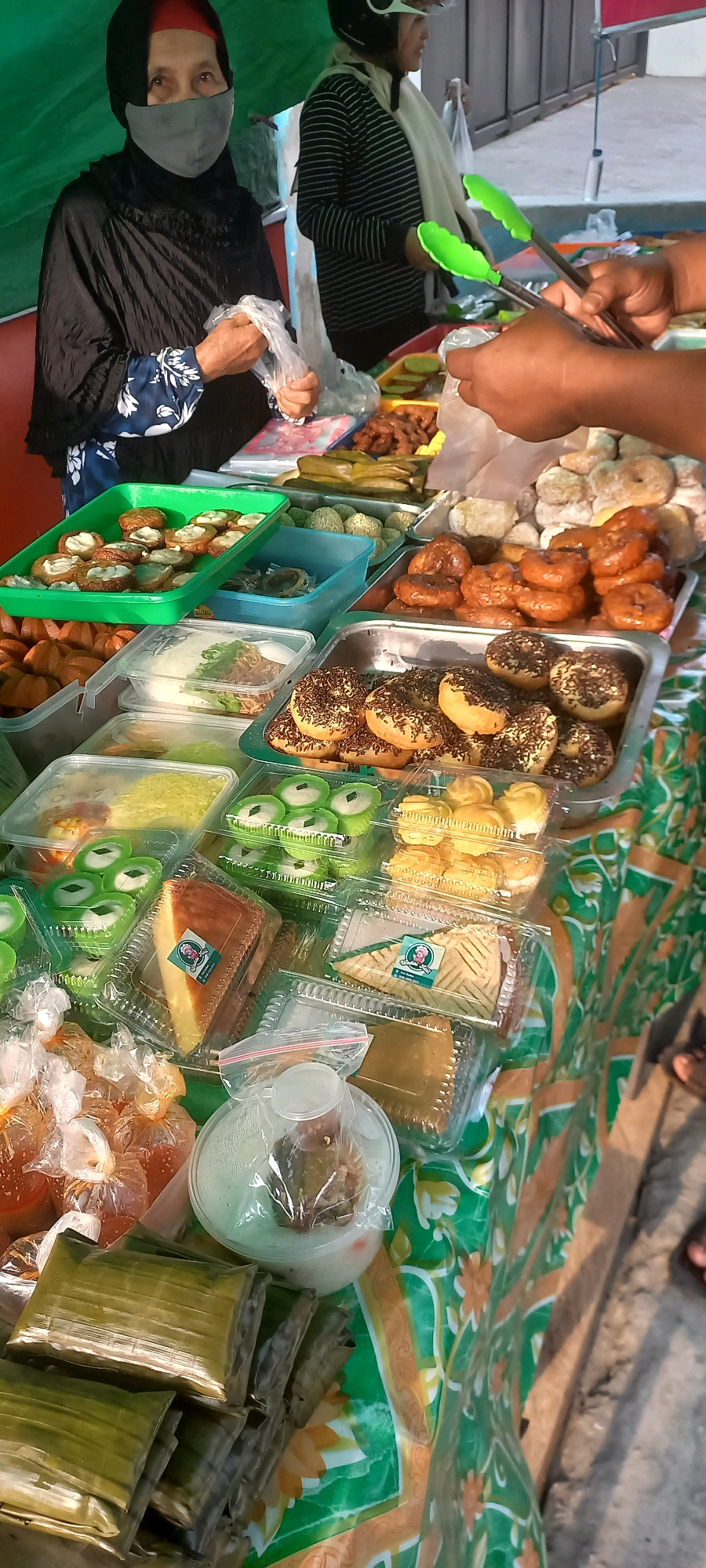 A street food stall is set up in Melayu, Indonesia. The stall is covered with a green and orange floral-patterned cloth. A woman wearing a black hijab and a grey mask stands behind the stall, holding a plastic bag. In front of her, various food items are displayed in plastic containers and trays. These include donuts topped with chocolate sprinkles, cream puffs, green pastries with white filling, a slice of cake, and what appear to be savory snacks wrapped in banana leaves. A person's hands are visible in the foreground, using green tongs to select food. The background shows a wall and a street.