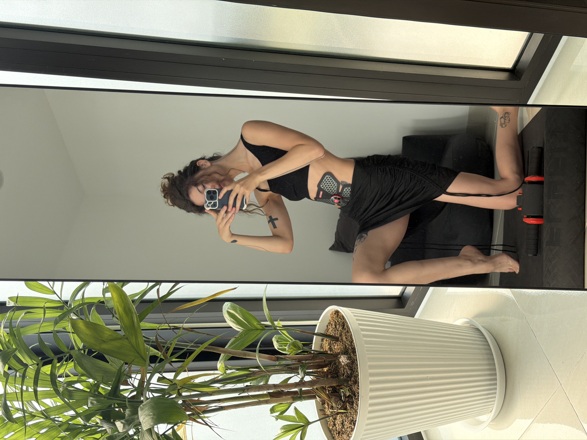 A young woman is visible in a mirror reflection. She is wearing a black sports bra and a black skirt, and she is holding a smartphone in her hands. She appears to be taking a selfie. A black EMS abdominal muscle trainer is attached to her abdomen. Tattoos are visible on her arms.
In the foreground, there is a houseplant in a white ridged pot. The plant's leaves are green and lush. The floor appears to be tiled. The image is taken inside what appears to be a modern building, possibly in Đà Nẵng, Vietnam, given the prompt. The specific location is not identifiable from the image. There is also an ab roller visible.