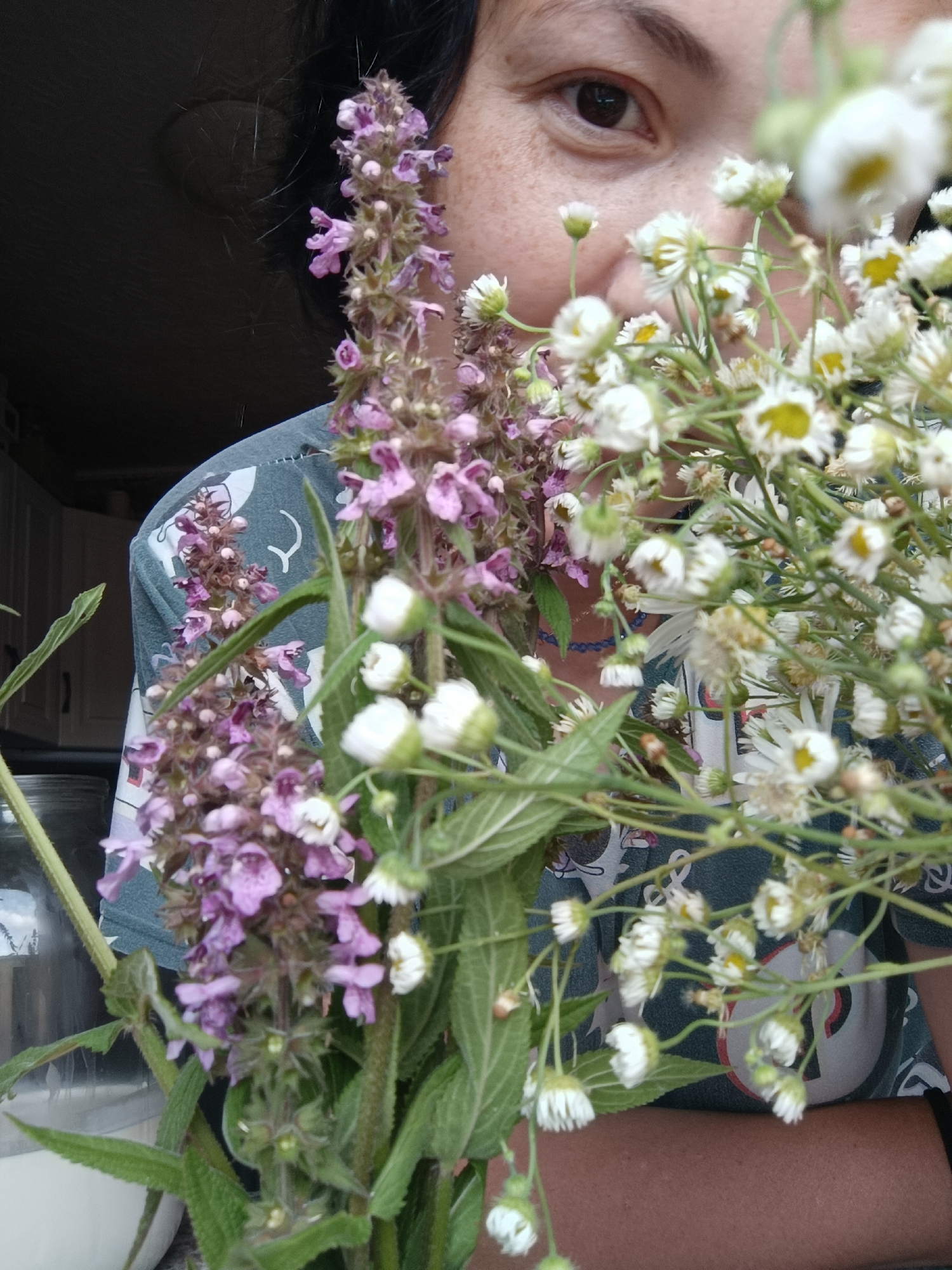 A close-up shot shows a woman from the shoulders up, with a bouquet of wildflowers held in front of her. She has dark hair and is looking towards the viewer with a slight smile. The flowers in the foreground are a mix of tall purple spires and smaller white daisies with yellow centers. The woman is wearing a dark teal t-shirt with a white graphic design. In the background, hints of a kitchen environment are visible, including cabinets and a glass jar. The lighting suggests an indoor setting, possibly near a window, with natural light illuminating the scene. The image does not contain any information related to the city of Karsha, Russia.