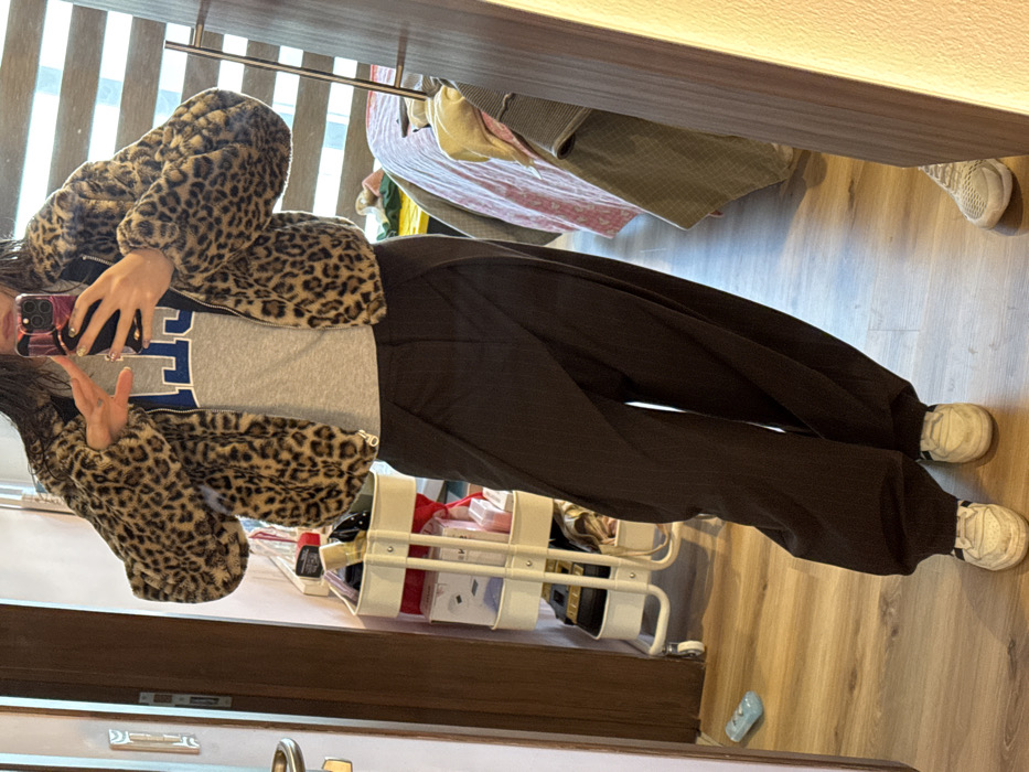 A young woman is seen from the torso down, wearing a gray T-shirt with blue lettering under a leopard-print jacket. She is also wearing dark brown, wide-leg pants. She is wearing white sneakers. She is holding a smartphone up as though taking a selfie in a mirror. Behind her, there is a metal shelf with items on it and some articles of clothing are hanging on a rack above her.