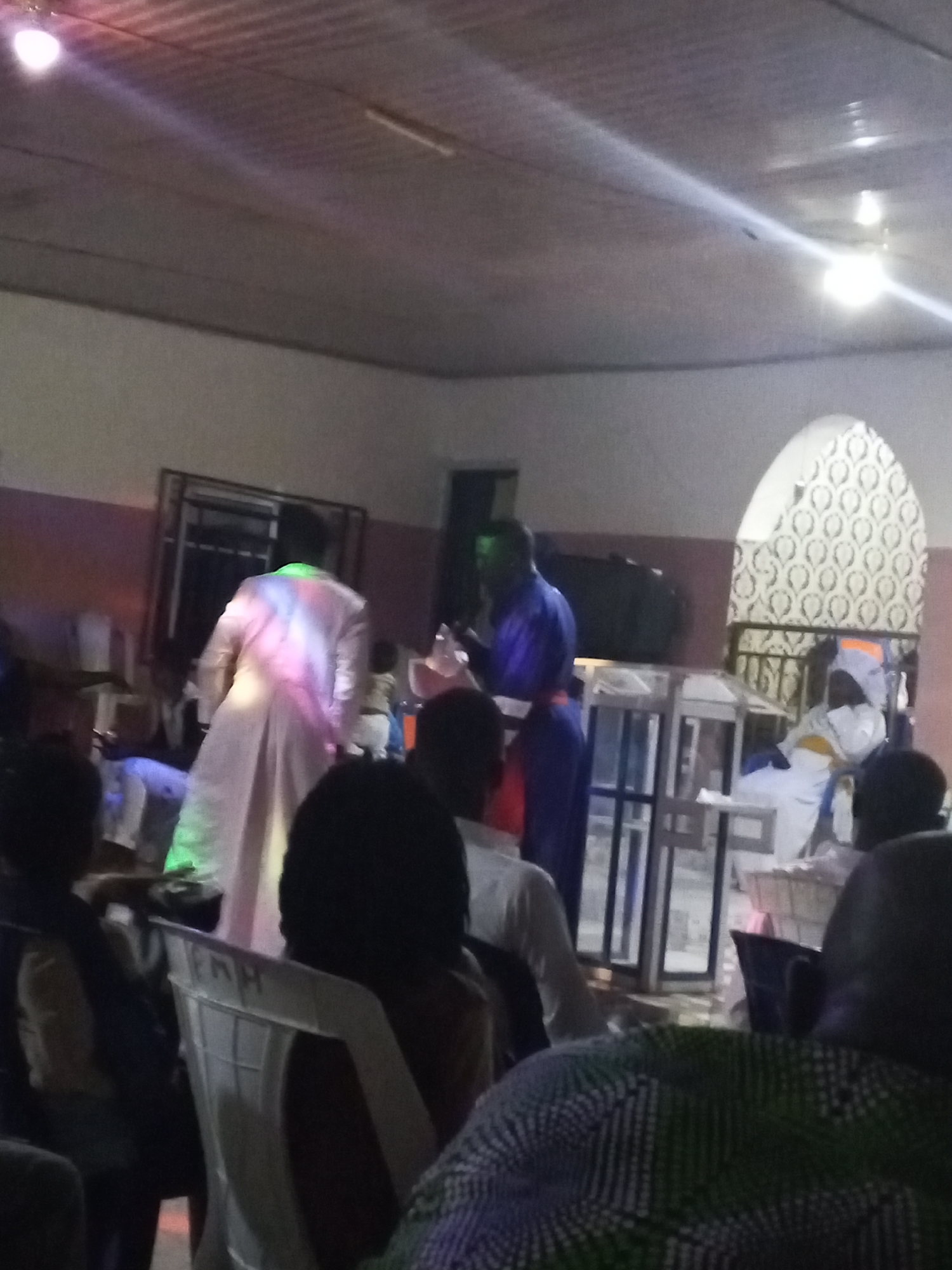 In a dimly lit indoor space, possibly a church in Lokoja, Nigeria, several individuals are gathered. A man in a long blue robe stands near a metal structure resembling a pulpit or lectern, while another man, wearing a white robe with green and pink lighting effects, walks away from the lectern. The room contains plastic chairs and what appears to be an archway decorated with patterns in the background. The lighting is poor, and the image is somewhat blurry.