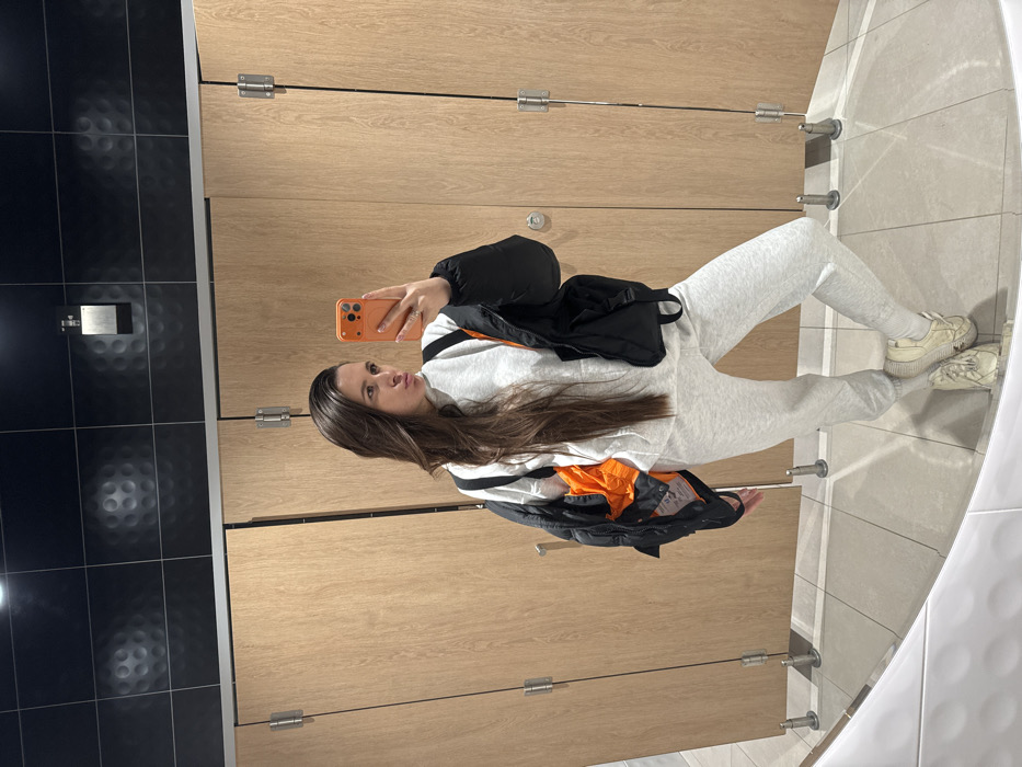 A woman takes a mirror selfie in a public restroom. She wears a gray sweatsuit, a black puffer jacket, and cream-colored sneakers. Her long, brown hair is down. She holds an orange phone case while facing the mirror. The bathroom has black and light wood paneled walls, chrome fixtures, and light tile flooring. A curved mirror reflects her image.