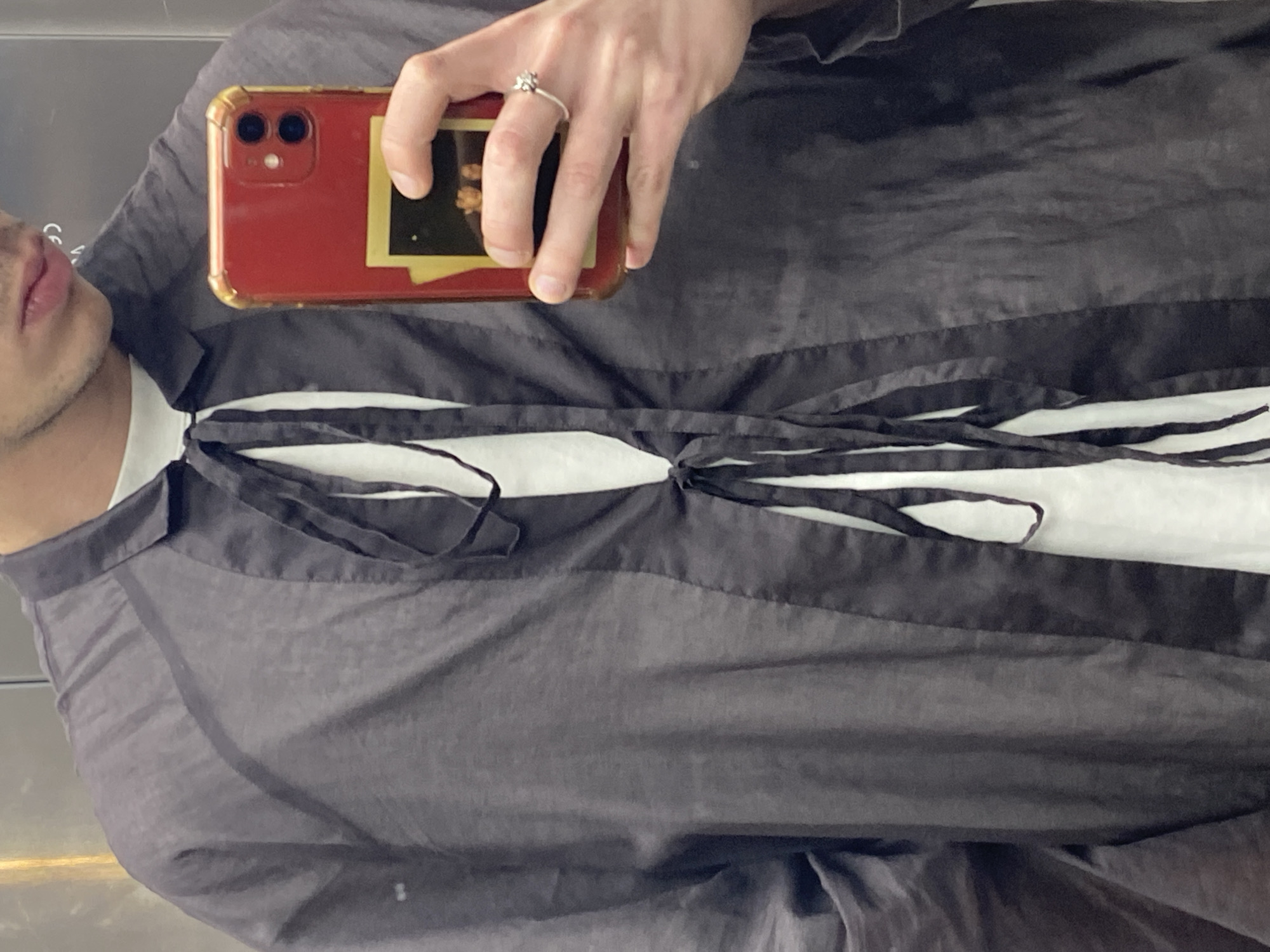 A person is holding a red smartphone in front of their chest. The phone has a snapshot photo attached to it, and the person is wearing a silver ring on their finger. The person is wearing a white shirt under a semi-transparent gray or black top with tie closures at the neckline. The image is oriented sideways, and the location is not determinable from the image.