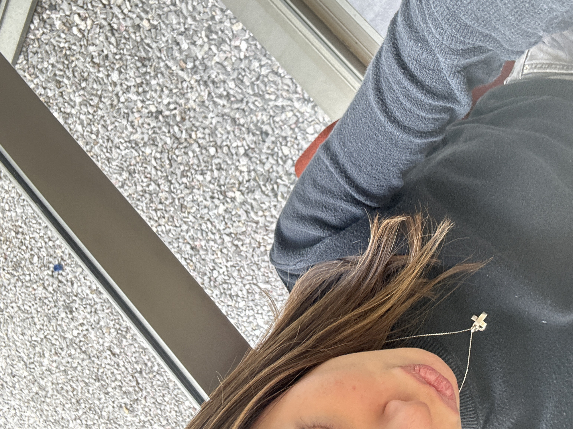 This close-up, diagonally oriented image features the lower part of a person's face and upper torso against a textured background. The subject has long, brown hair that falls across their face and chest. They are wearing a dark grey or black long-sleeved knitted sweater. A silver-colored necklace with a small "X" shaped pendant is visible, resting on the sweater. The shot includes the person's lips and chin, with no discernible emotion.
The background consists of a light grey, granular surface, resembling fine gravel or aggregate pavement. A dark grey, rectangular metallic beam or rail runs diagonally across the scene, appearing to be part of a structure like a balcony railing or a modern building element. Another, thinner metallic line is visible further behind it. The lighting is bright and even, suggesting daytime conditions without direct sunlight. The setting appears to be outdoors or semi-outdoors. There is no visible text within the image.