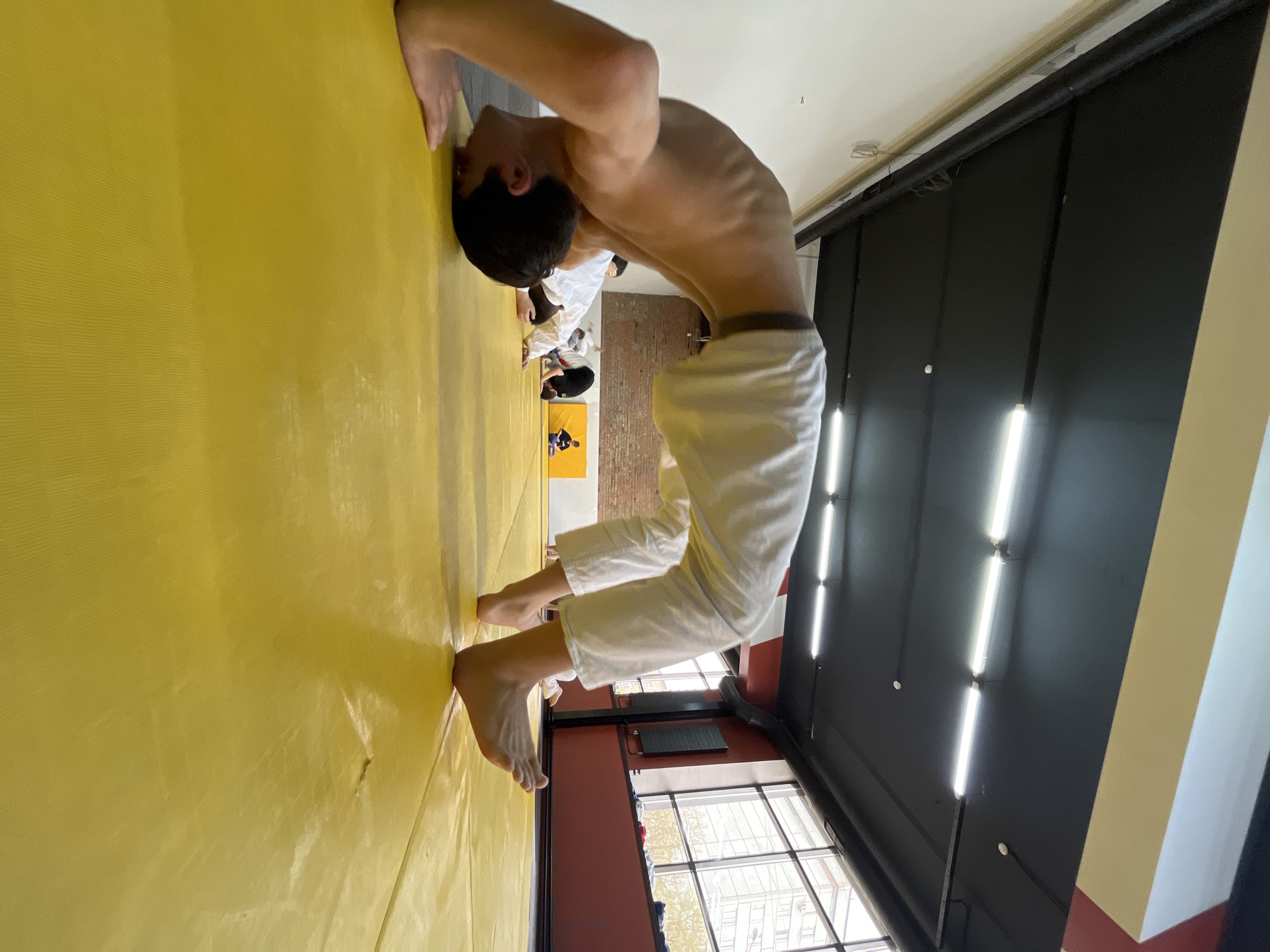 A young man is performing a bridge exercise against a yellow wall in a room with fluorescent lighting. He's barefoot and wearing white pants. Other individuals, dressed similarly, are visible in the background. A window is visible near the bottom of the frame, and the room appears to be a martial arts dojo. Based on the image's location metadata, this scene is in Krasnoyarsk, Russia.