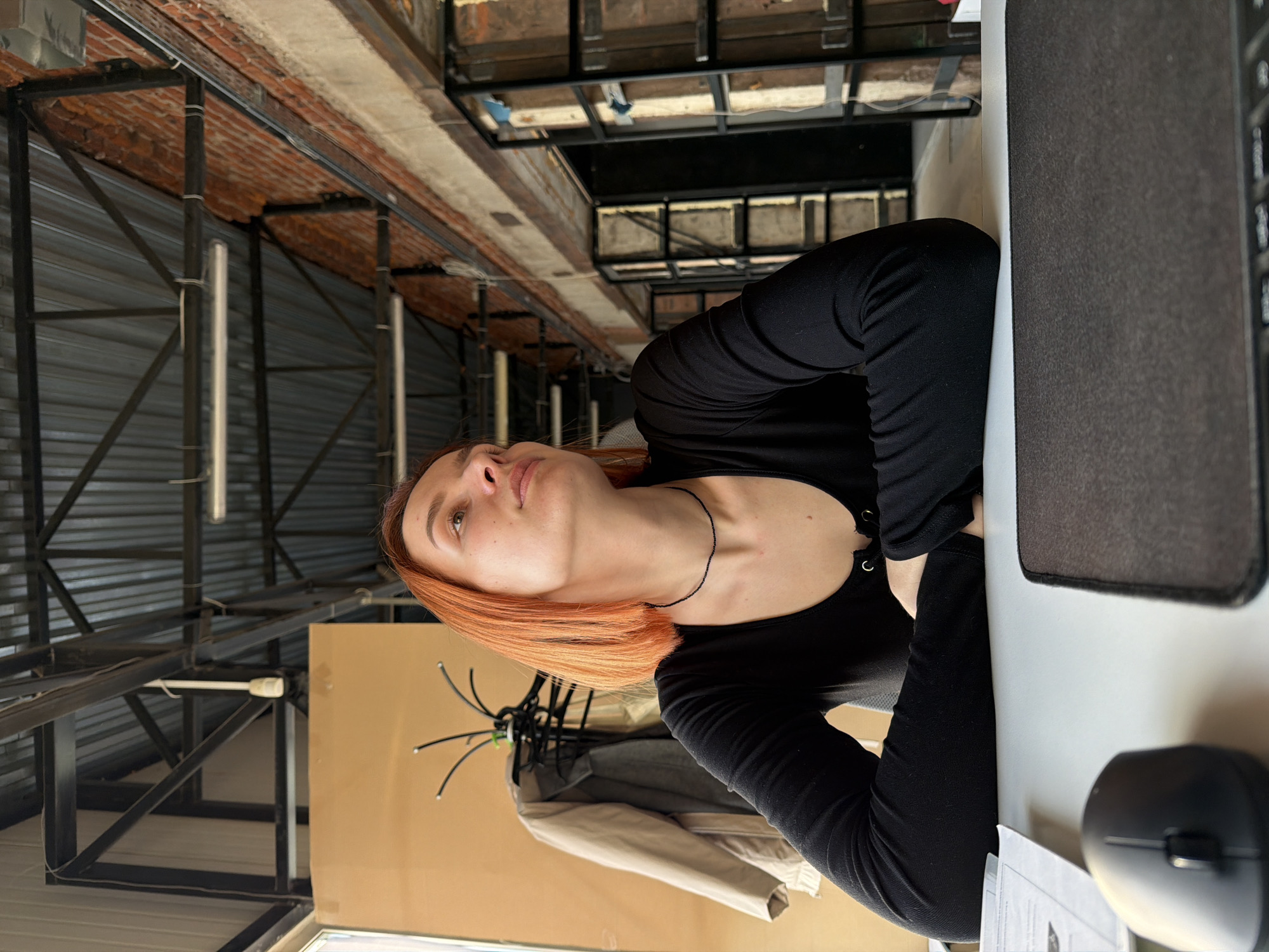 The image depicts a woman with reddish-orange hair, wearing a black long-sleeved top, sitting at a desk with a computer, mouse, and papers. Behind her, a light-colored jacket hangs on a rack against a wall. The setting appears to be inside a building with an industrial-style ceiling featuring metal beams and corrugated metal sheets. The location is potentially within the city of Chaikovsky, Russia, based on the prompt's requirement.