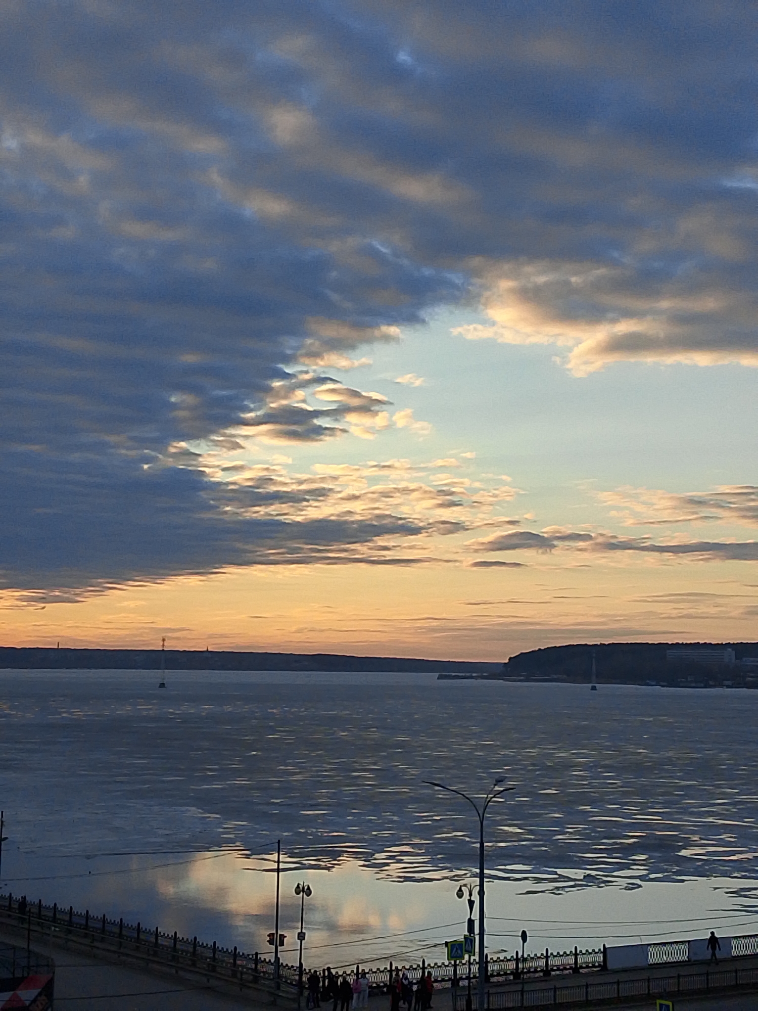 The image depicts a cityscape scene in Izhevsk, Russia, at dusk or dawn, with a partially frozen river reflecting the colorful sky. Above the horizon is a cloudscape displaying a mix of blue, grey, and orange hues. Below this sky is a wide expanse of water, likely the Izhevsk Pond, where ice floes float near the surface, reflecting light. In the foreground is a paved area with a decorative railing, lampposts, and streetlights, indicating it is a promenade or waterfront walkway. Several people are visible on the walkway, appearing as small figures walking along or gathering in groups. In the distance across the water are darker landmasses, likely the opposite bank of the pond, where buildings and vegetation can be indistinctly observed. The overall ambiance suggests a tranquil, cold, and urban setting.