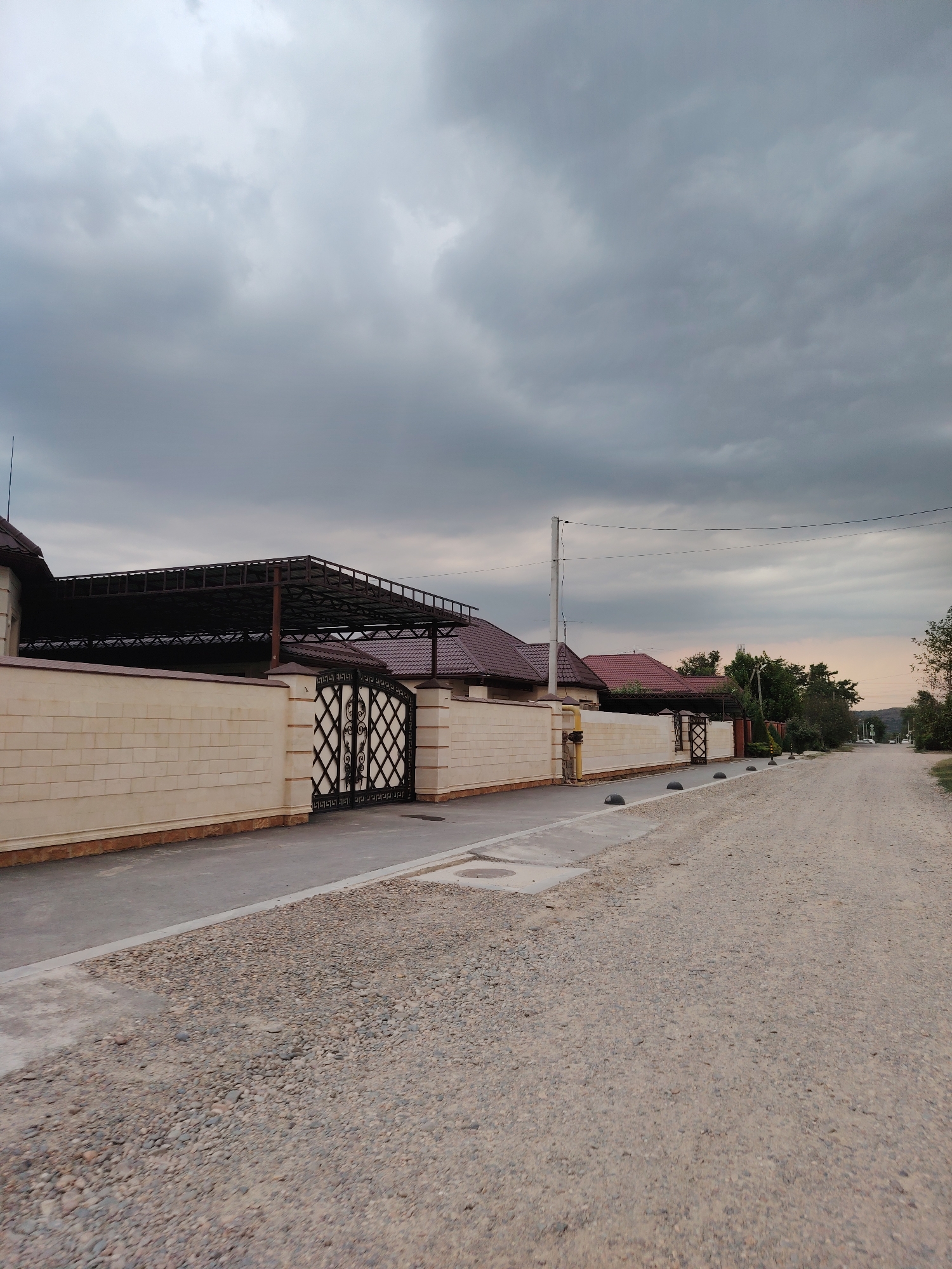 The image shows a street in Volnoye, Russia. The street is unpaved and covered in gravel. On the left side of the street is a row of houses with red tile roofs. The houses are separated by a low, beige brick wall. There is a black metal gate in front of one of the houses. The sky is overcast and cloudy. The overall impression is of a rural or suburban setting.