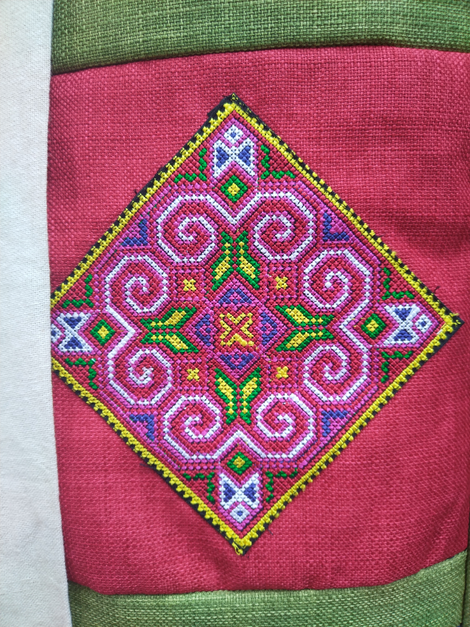 The image depicts a piece of handmade textile art. A red fabric is the background, on which a symmetrical, diamond-shaped embroidered pattern is centered. The embroidery features swirls and geometric designs in white, blue, green, yellow, pink, and black. The outer edge of the diamond is bordered with yellow and black stitching. The textile art is framed with green woven fabric above and below, and with white fabric to the side. The image suggests a product of Sa Pa Ward, Vietnam, known for its traditional handmade textiles.
