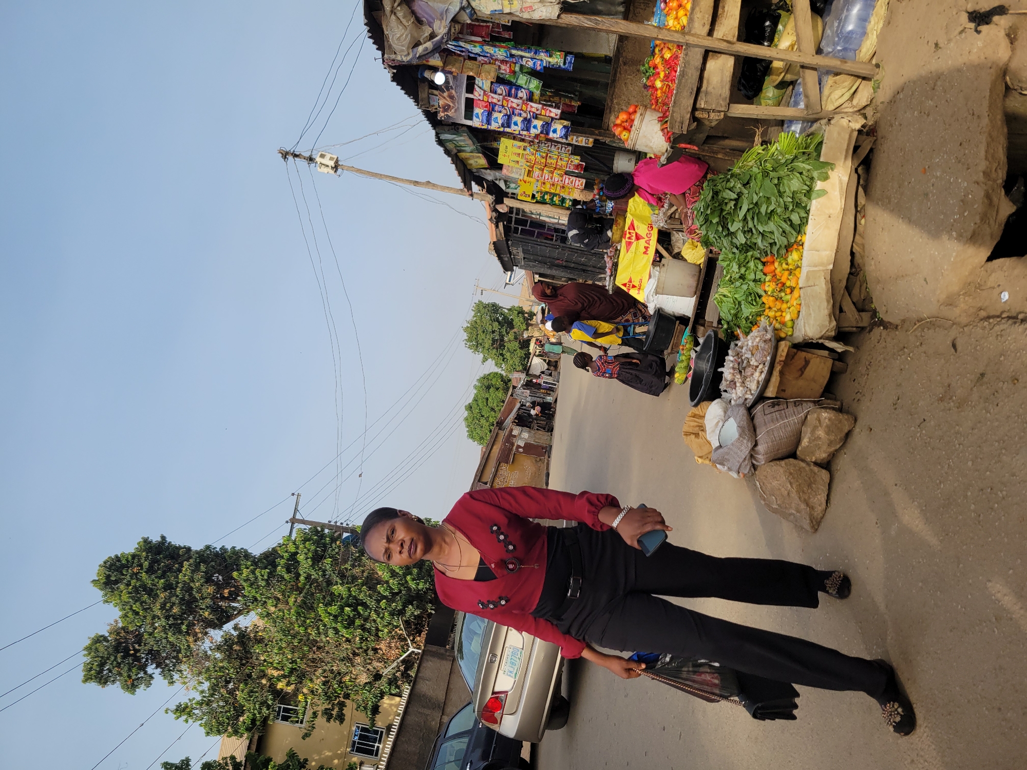The image shows a street scene in Jos, Nigeria. A woman in a red, long-sleeved top and black pants stands in the foreground. She holds a mobile phone in her right hand and a purse in her left. Behind her are parked cars, and a building partially obscured by trees is visible to the left. To her right and further into the background, a roadside market is set up. Vendors are seated beside stalls selling produce like green leafy vegetables, tomatoes, and other items. People are seen walking along the street. Overhead, electrical wires connect to a pole. The sky is clear and light blue.