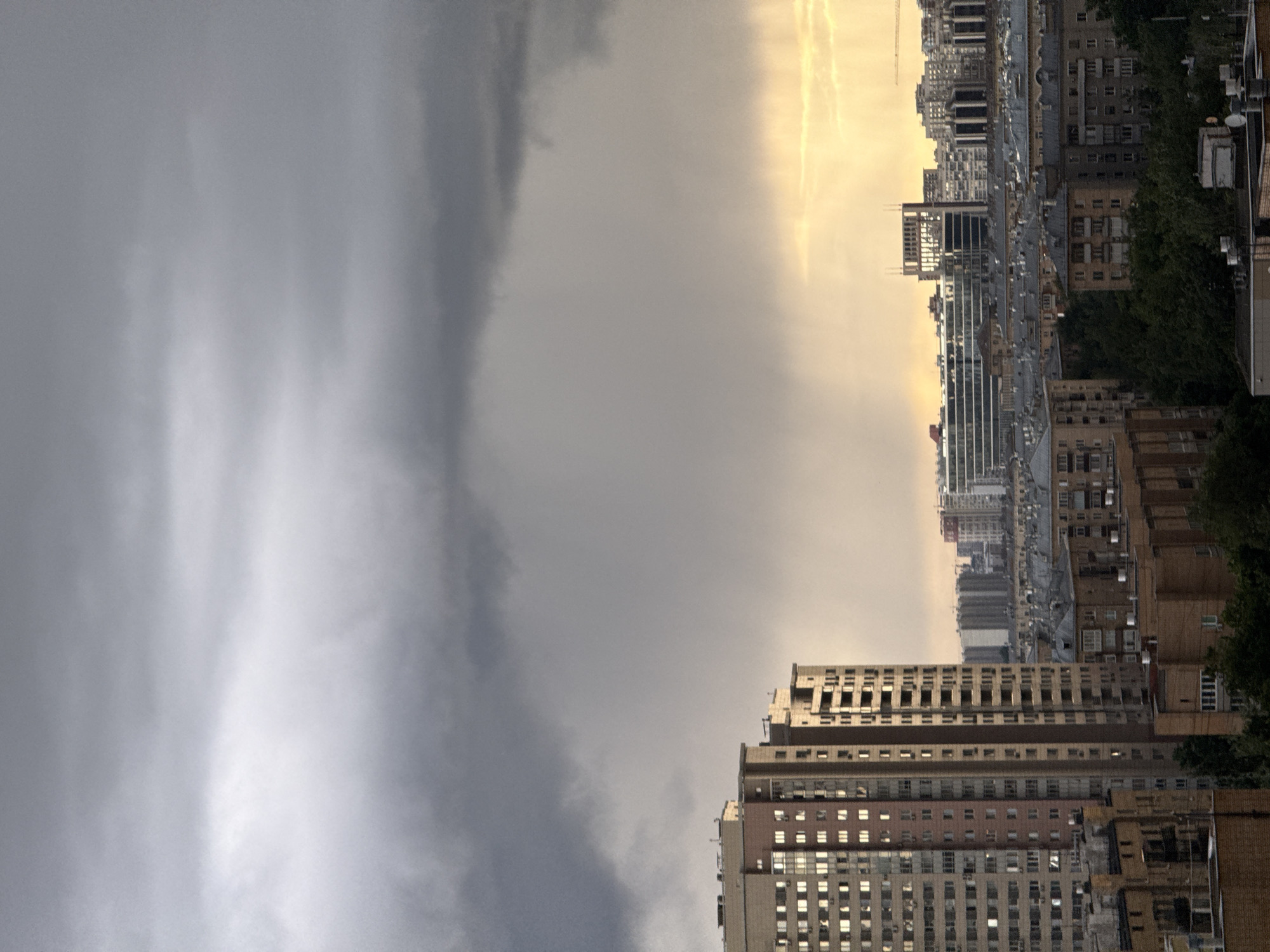 The image shows a cityscape, likely Moscow, Russia, bisected by contrasting weather conditions. The right side is illuminated with a bright, golden light filtering through a hazy sky, highlighting residential and commercial buildings of varying heights. These buildings appear to be constructed with brick and concrete and show signs of age, with many windows and some greenery interspersed. The left side of the image is dominated by a dark, ominous storm cloud. The cloud cover is dense and heavy, suggesting an impending downpour. The contrast between the sunlit city and the approaching storm is stark. The composition is split roughly in half, emphasizing the dramatic weather event. The image is oriented vertically, suggesting it was taken from a high vantage point or with a phone camera.