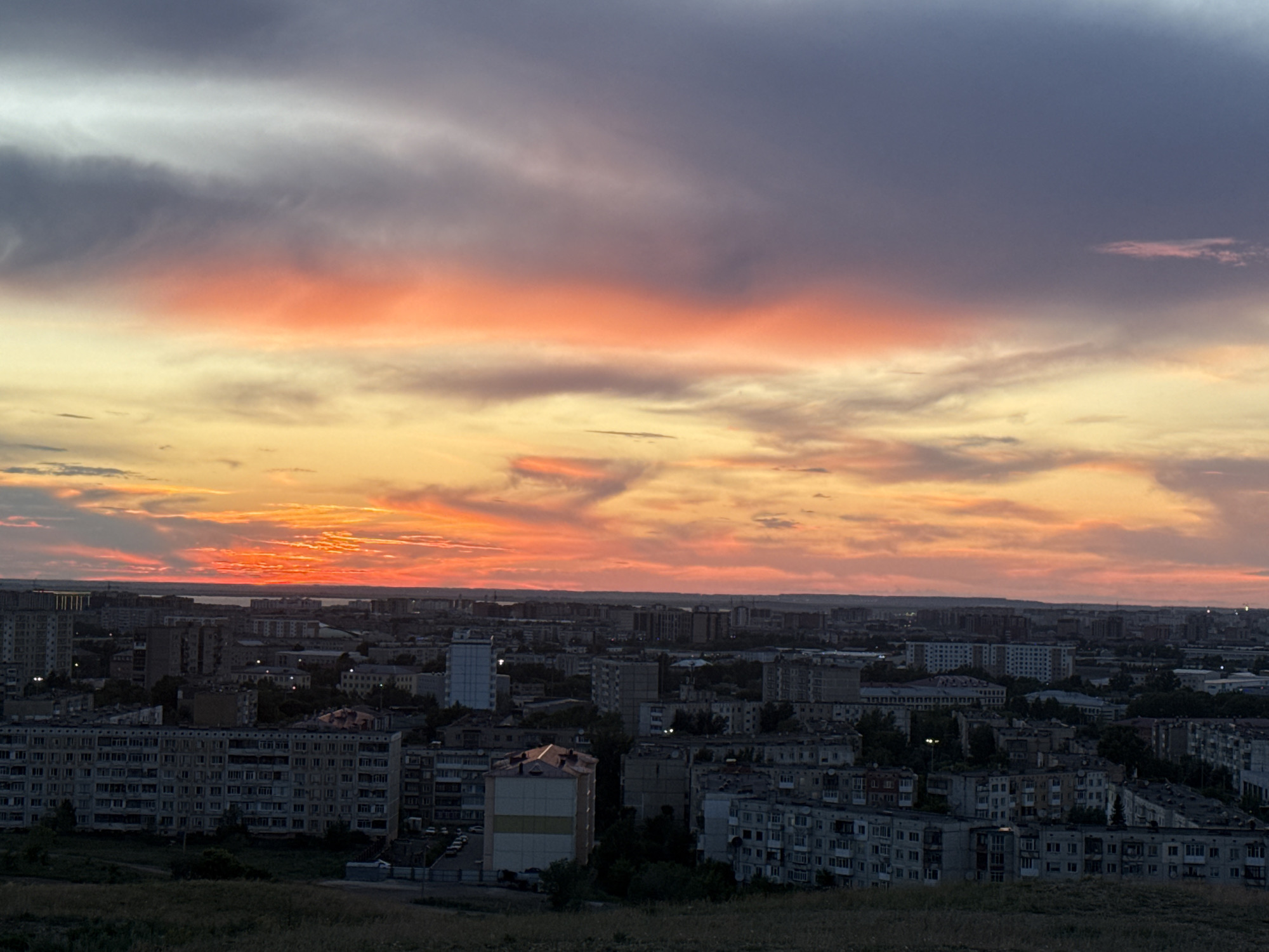 This is a medium-distance, high-angle shot of a city skyline at sunset. The sky is dominated by vibrant orange and yellow hues, with streaks of gray and purple clouds. Below the sky, a dense urban landscape stretches to the horizon. The buildings are primarily apartment blocks of varying heights, many appearing to be Soviet-era architecture with repetitive window patterns. Some taller, more modern buildings are also visible. The foreground features a grassy hill with dry, muted vegetation. The overall impression is of a residential area within a city, bathed in the dramatic light of a setting sun. The image is likely taken from a higher vantage point overlooking the city, possibly a hill or elevated area within Kokshetau, Kazakhstan, given the architectural style and landscape characteristics that align with descriptions of the region. No people are visible in the image. The scene is static, focusing on the cityscape and the sky.