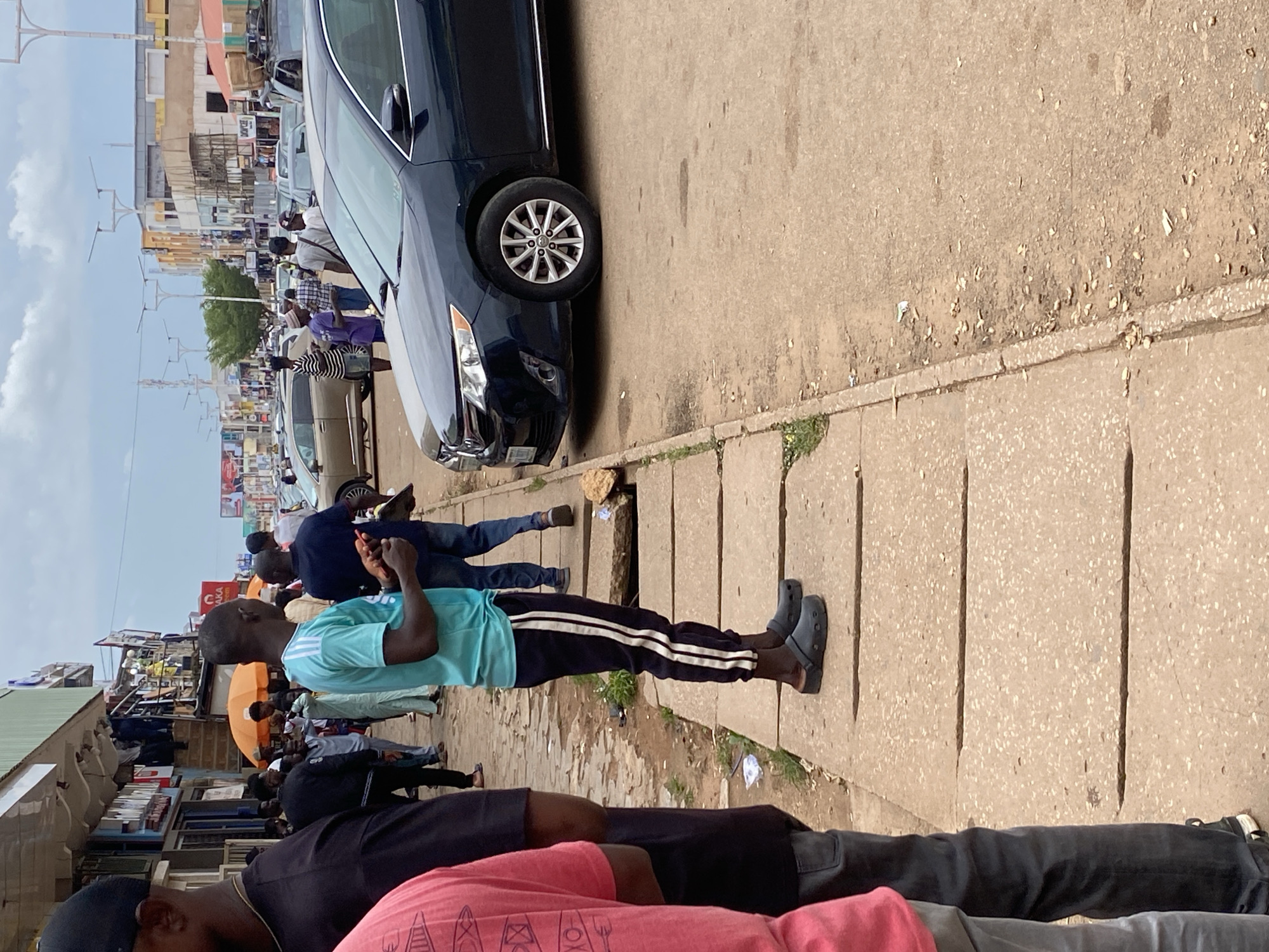 A medium shot captures a street scene with pedestrians and vehicles in an urban environment. In the foreground, on a concrete sidewalk, a man in a teal t-shirt and dark pants with white stripes stands facing right, holding a small red object in his right hand and a dark object in his left. He is wearing dark clogs. To his left, a portion of another person's pink shirt and dark pants is visible. Further down the sidewalk, more individuals are seen walking and standing. In the middle ground, several vehicles, including a dark sedan, are on the road. Shops with awnings and signs line the street. The background shows buildings and streetlights under a cloudy sky. The overall scene depicts a bustling commercial area.