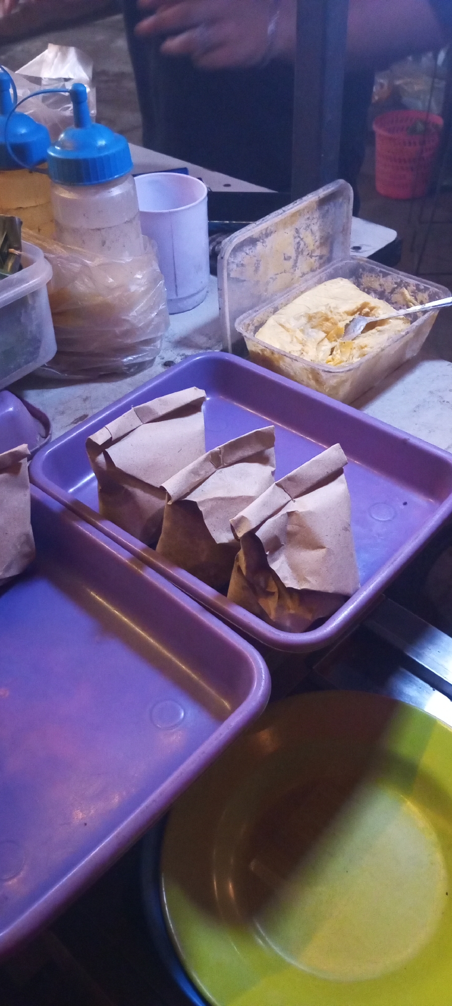 In Setu, Indonesia, a close-up, slightly blurry, eye-level shot captures a food stall scene. In the foreground, a purple plastic tray holds three small, folded brown paper bags, likely containing food items. To the left of this tray, another purple plastic tray is visible, mostly empty. Behind the paper bags, a clear plastic container holds a yellow, somewhat lumpy substance, with a metal spoon resting in it. Further back, a white plastic cup is positioned next to a clear plastic bottle with a blue cap, suggesting it might be a sauce dispenser. A white plastic bag filled with an unknown substance is partially visible to the left of the bottle. The background is dimly lit, with a glimpse of a person's hands and arms, indicating activity at the stall. A red plastic basket is also visible in the background. The overall impression is one of a busy, informal food service environment.