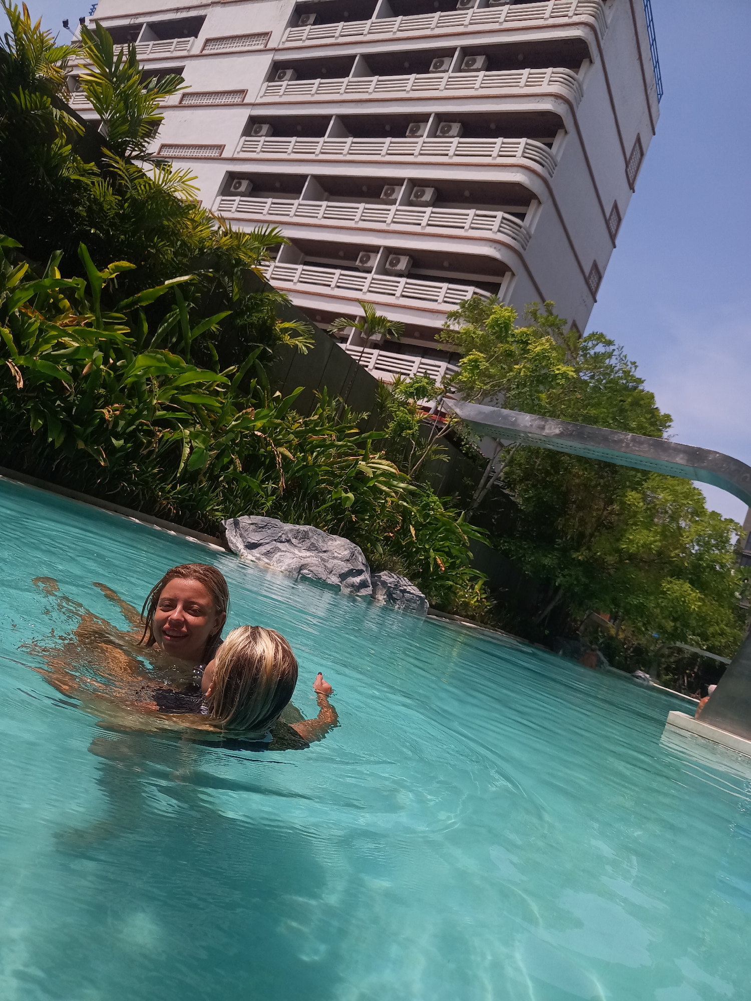 The image shows two light-skinned women swimming in an outdoor pool, likely in a hotel setting. The pool water is clear and turquoise. Lush green vegetation borders the pool area. In the background is a tall, white building with multiple floors and balconies, each equipped with an air conditioning unit. A silver, arched structure hovers over the pool. The location is Hua Hin City Municipality, Thailand.