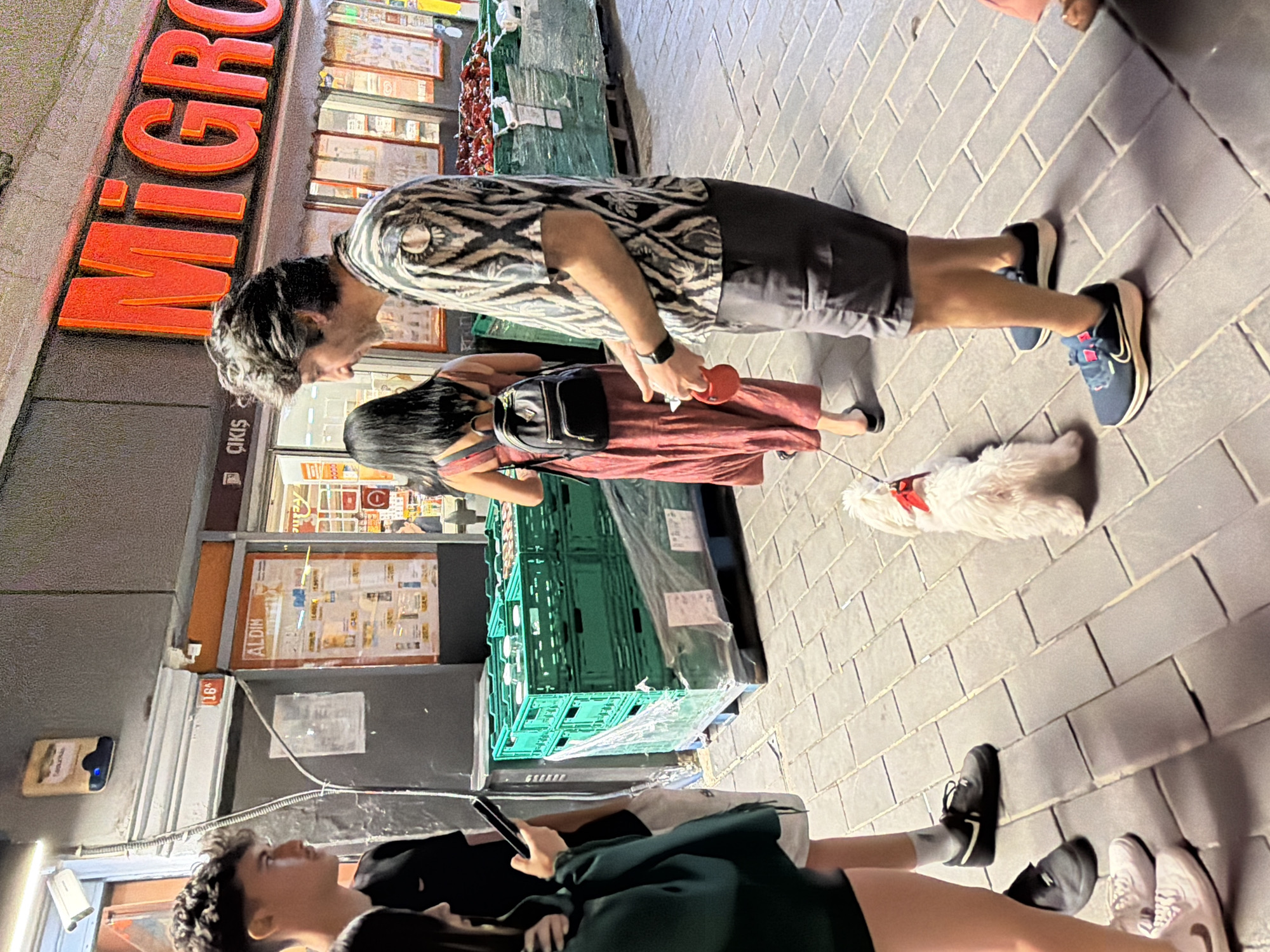 The image depicts a street scene in Kadıköy, Turkey, likely in the evening, with a prominent "Migros" supermarket sign illuminated in orange neon. In the foreground, a man with dark, curly hair wearing a patterned short-sleeved shirt and grey shorts holds a red dog leash attached to a small, fluffy white dog with a red collar. The dog is facing away from the camera. To the left of the man, a woman in a long, reddish-pink dress is visible from the back, wearing a backpack and walking away from the camera. Beside her, a young person with dark, curly hair, wearing a black t-shirt, stands with their back to the camera, looking to their left and holding a dark smartphone. Further to the left, partially visible, is another person wearing a dark green hooded garment and black shorts. Behind the main subjects, green plastic crates filled with goods are stacked on pallets. In the background, the entrance to the Migros store is visible, with displays of produce, including red items that appear to be tomatoes, on the right side. Adjacent to the Migros, a store with "ALDI" signage is also present. The ground is paved with grey rectangular tiles. A security camera is mounted on the building wall above and to the left of the "ALDI" sign.