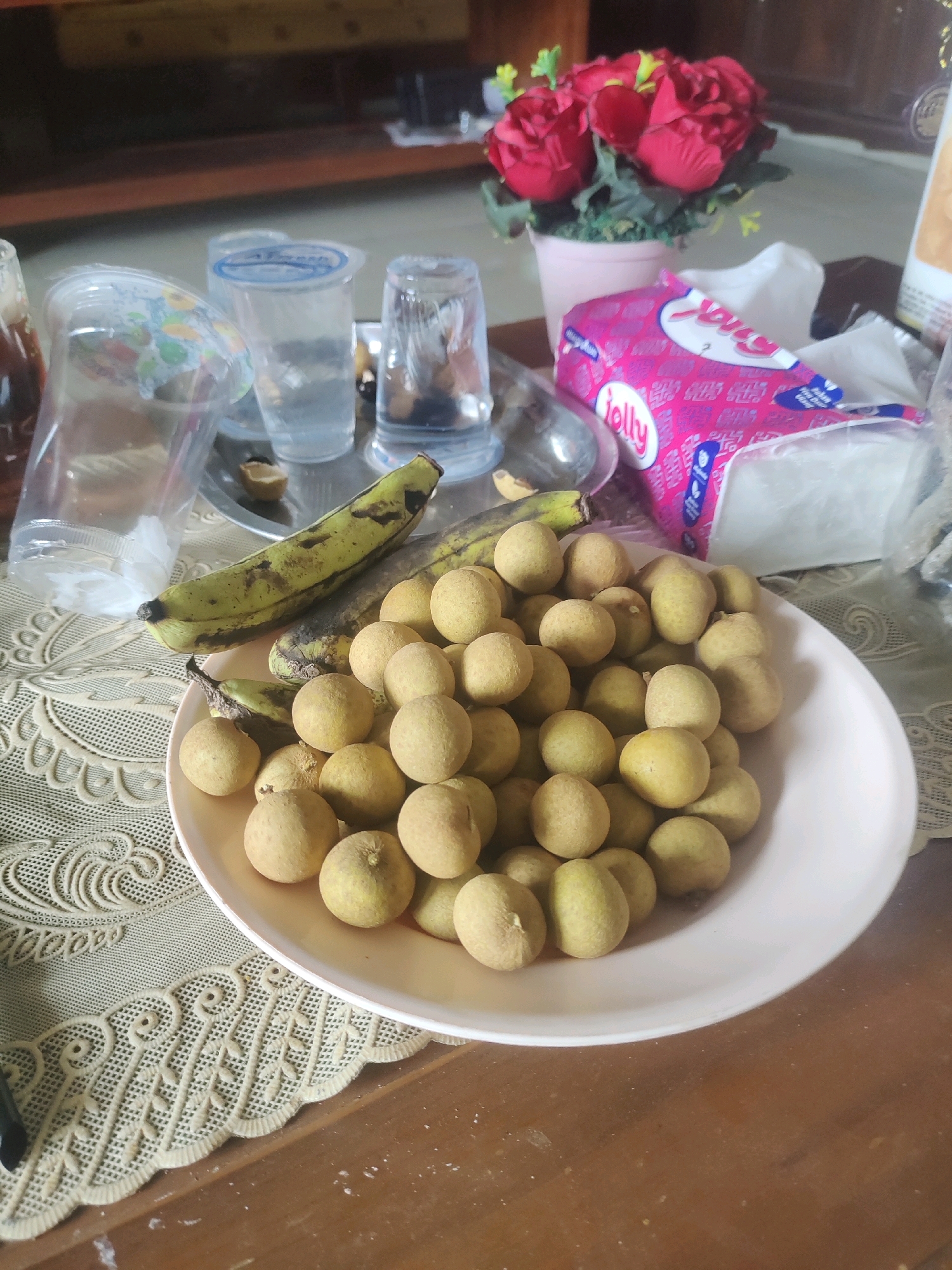 A close-up shot, taken from a slightly high angle, shows a collection of longans and bananas arranged on a table covered with a textured beige tablecloth. The longans are piled high in a light pink bowl, with some spilling over the edge. They are round and have a light brown, slightly fuzzy skin. Two bananas, one mostly yellow with brown spots and the other greener with brown marks, are placed next to the bowl.
In the background, several clear plastic cups and a glass with water are visible, along with a pink package of tissues that has the word "Jolly" printed on it. Behind the tissues, a small potted plant with red artificial roses and green leaves stands out. The table appears to be made of wood, and there are some white specks scattered on its surface. The overall scene suggests a casual setting, possibly a home or a market stall in Rejowinangun, Indonesia, where these fruits are commonly found.