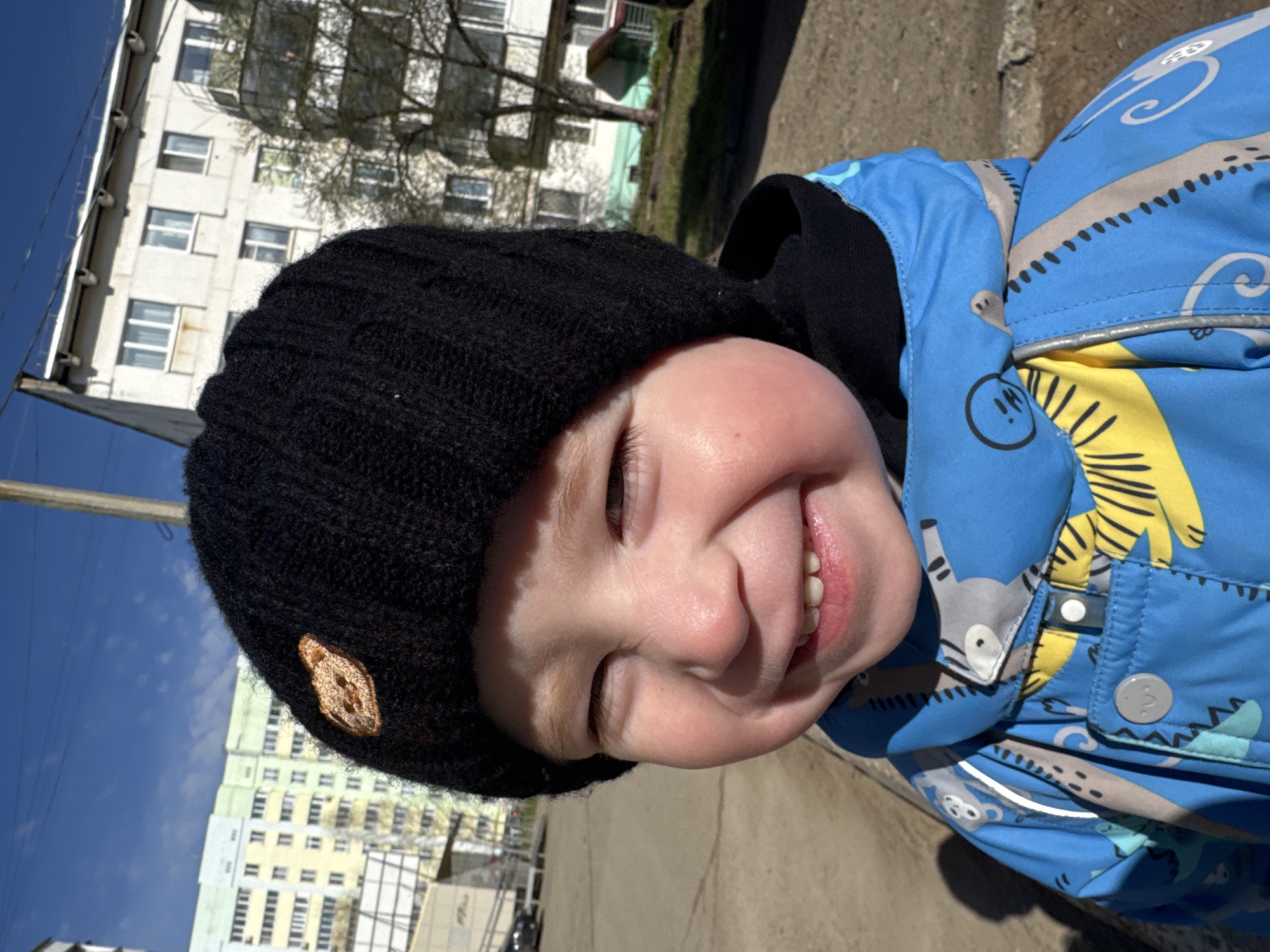 A young child, likely a toddler, is captured in a close-up outdoor portrait, smiling joyfully and looking directly at the camera with a playful expression, including a slightly extended tongue. The image is rotated, but depicting the child in an upright orientation, they are wearing a black knit beanie, which features a small, light-brown, textured patch resembling an animal head on its side. Over a dark undershirt, the child wears a bright blue jacket adorned with a lively pattern of cartoon animals, possibly raccoons and monkeys, alongside abstract swirls in shades of gray and yellow.
The background reveals a typical urban or suburban residential setting. Multi-story apartment buildings, likely concrete or stucco, with numerous windows are visible under a clear, bright blue sky with a few wispy clouds. Bare trees with visible branches suggest late winter or early spring. The ground beneath appears to be a paved surface like asphalt or concrete. A utility pole can be seen on the left side of the frame. The scene is bathed in bright daylight, indicating sunny weather. The overall mood is cheerful and innocent. There is no discernible text visible in the image. The location context is Chaikovsky, Russia.