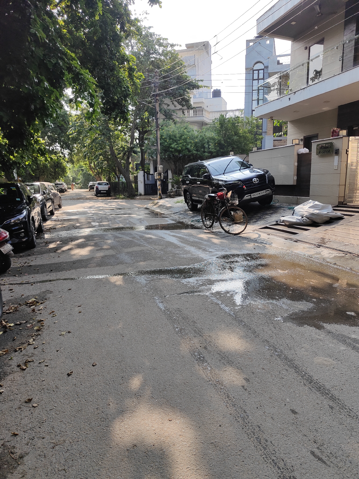 In a residential street in Gurgaon, India, a black SUV is parked in front of a modern house. Water is pooled on the asphalt road, reflecting the sunlight and creating a glistening effect. Tire tracks are visible on the wet surface. A bicycle with a basket is positioned on the side of the road near the SUV. Trees line the street, casting shadows on the pavement. Other vehicles are parked further down the road. The sky is bright, suggesting daytime.
