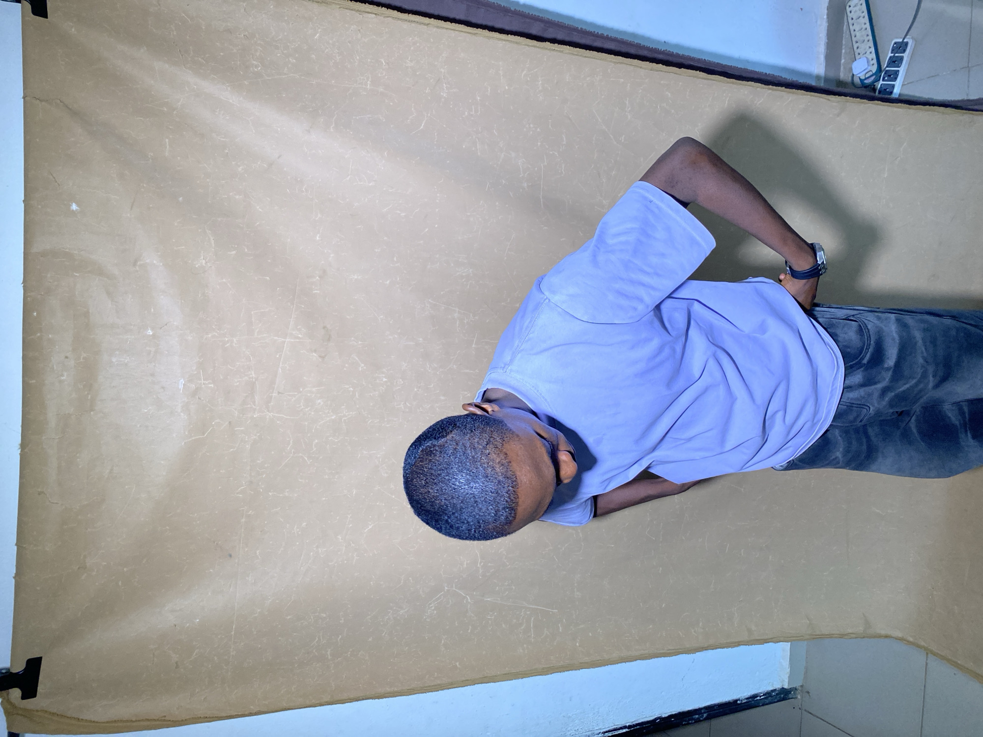 A young Black male is depicted in an indoor setting, possibly a makeshift photography studio in Bukuru, Nigeria. He is standing upright, wearing a light blue short-sleeved t-shirt and dark pants, with a dark-strapped wristwatch on his left wrist. His head is shaved with a short buzz cut, and he is looking downwards and slightly to his right, with his hands clasped behind his back. He appears to be posing or waiting.
Behind him, a light brown or beige seamless paper or fabric backdrop covers the wall and extends onto the floor. The backdrop shows visible wrinkles and textures, indicating wear or an imperfect setup. To the right of the subject, on a light blue wall, a white power strip with several outlets is visible, with some black and white cables plugged into it. A dark trim lines the bottom of the wall. The image is captured from a high-angle perspective, looking down at the subject and the immediate surroundings. The lighting appears soft and even. No discernible text is visible in the frame.