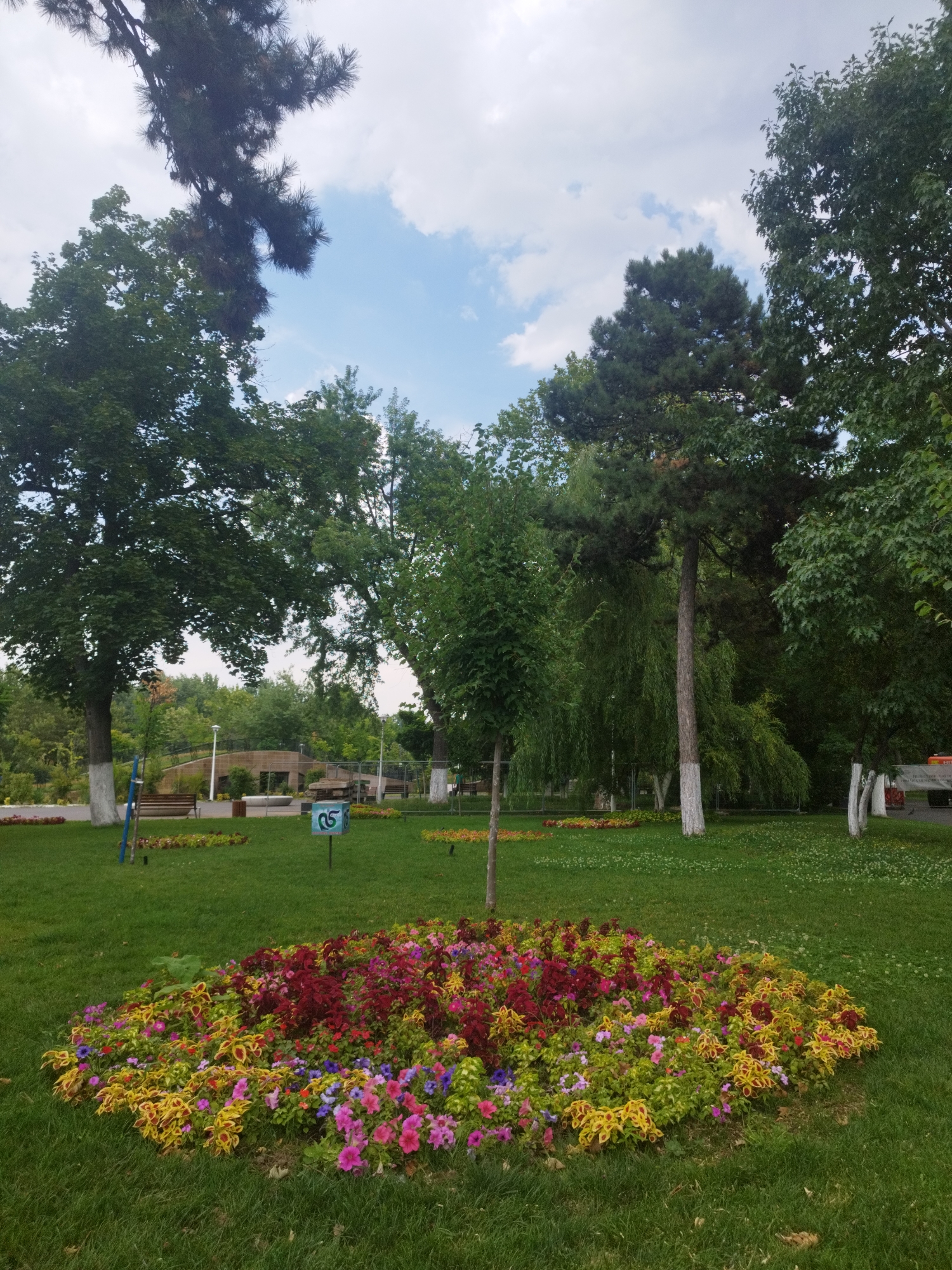 The image depicts a vibrant flowerbed in a park in Bucharest, Romania. The flowerbed is circular and filled with a variety of colorful flowers, including red, yellow, pink, and purple blooms. The flowers are densely planted, creating a rich tapestry of color. In the background, several trees with lush green foliage are visible, as well as a paved path and park benches. The sky is partly cloudy with patches of blue. A sign with the number "05" is visible on a post in the foreground.