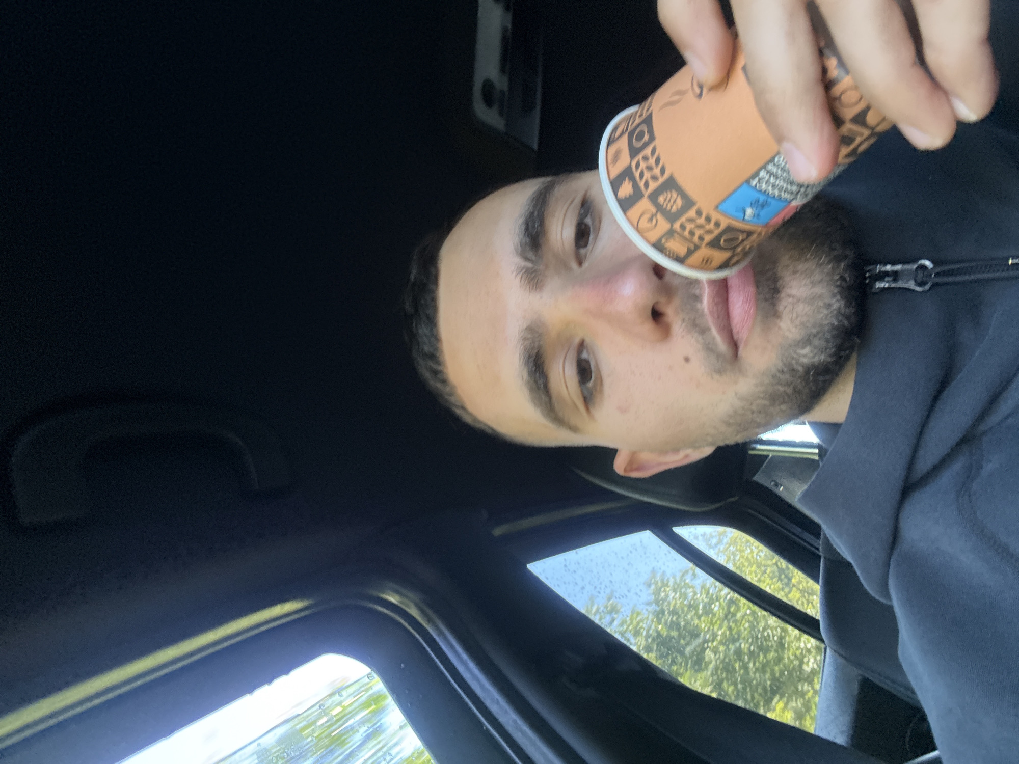 The image is a close-up, angled selfie of a young man taken inside a vehicle. The man, likely in his 20s or 30s, has short dark hair, distinct eyebrows, and a trimmed beard or stubble. He is wearing a dark hooded jacket with a prominent silver zipper. His face is angled towards the camera, with a neutral to slightly contemplative expression.
He holds an orange and brown paper cup with his right hand, positioning it near his mouth as if about to drink or having just taken a sip. The cup features various decorative patterns including coffee beans, mugs, and geometric shapes.
The setting is the interior of a car or similar vehicle, with the dark headliner filling much of the upper left portion of the frame. Through the windows on the right and bottom, bright green foliage from trees and glimpses of a bright sky are visible, indicating an outdoor, daytime scene. Small water droplets are noticeable on the window panes, suggesting recent rain or condensation. There is a grab handle or similar car interior feature visible on the left side of the frame. No legible text is visible on the cup or in the background. The location context is Хаджийско, Bulgaria.