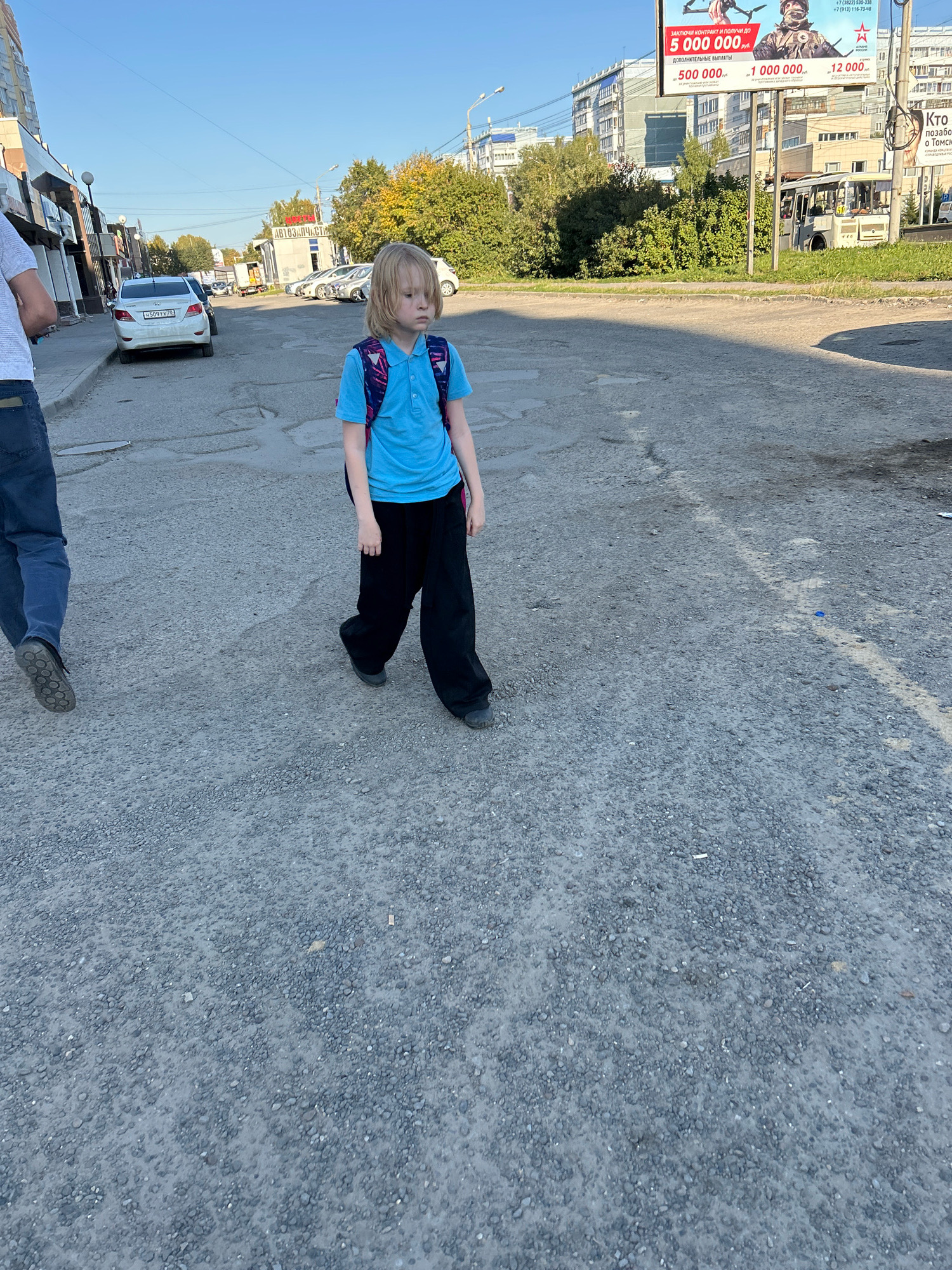 The image depicts a young boy with blonde hair, wearing a blue polo shirt and black pants, walking across a gravelly road in Tomsk, Russia. He has a blue and pink backpack on his shoulders. To the left, a person wearing a light-colored t-shirt and blue jeans is also walking away from the camera. In the background, there are buildings, trees, cars, and a bus. A billboard with Russian text and images is visible on the right side of the image. The sun is shining, casting shadows on the road.